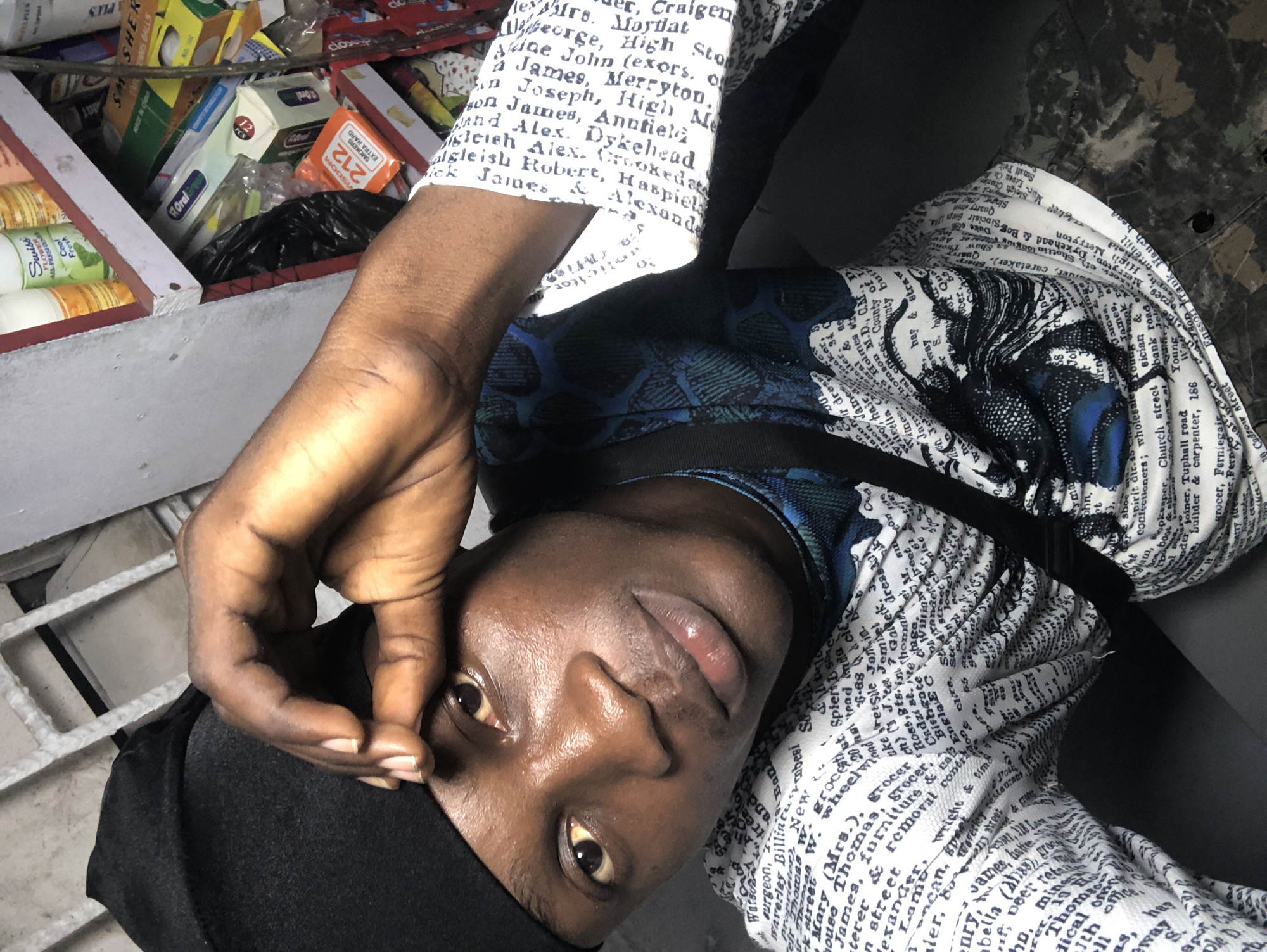 The image is a close-up selfie of a young Black male wearing a black skullcap and a white t-shirt with a repeating pattern of text. The text appears to be a list of names and professions, some of which are legible as "grocer", "builder & carpenter", and "street". He is looking towards the camera with his right hand raised to his forehead, obscuring part of his left eye. His left hand is resting on his chest. Behind him, a shelf is visible, stocked with various items including bottles of air freshener, boxes of toothpaste, and other retail products. The background suggests an indoor setting, possibly a shop in Ado, Nigeria, given the context. The lighting is somewhat dim, with shadows cast on the subject's face. The overall impression is casual and personal, a snapshot taken by the subject himself.