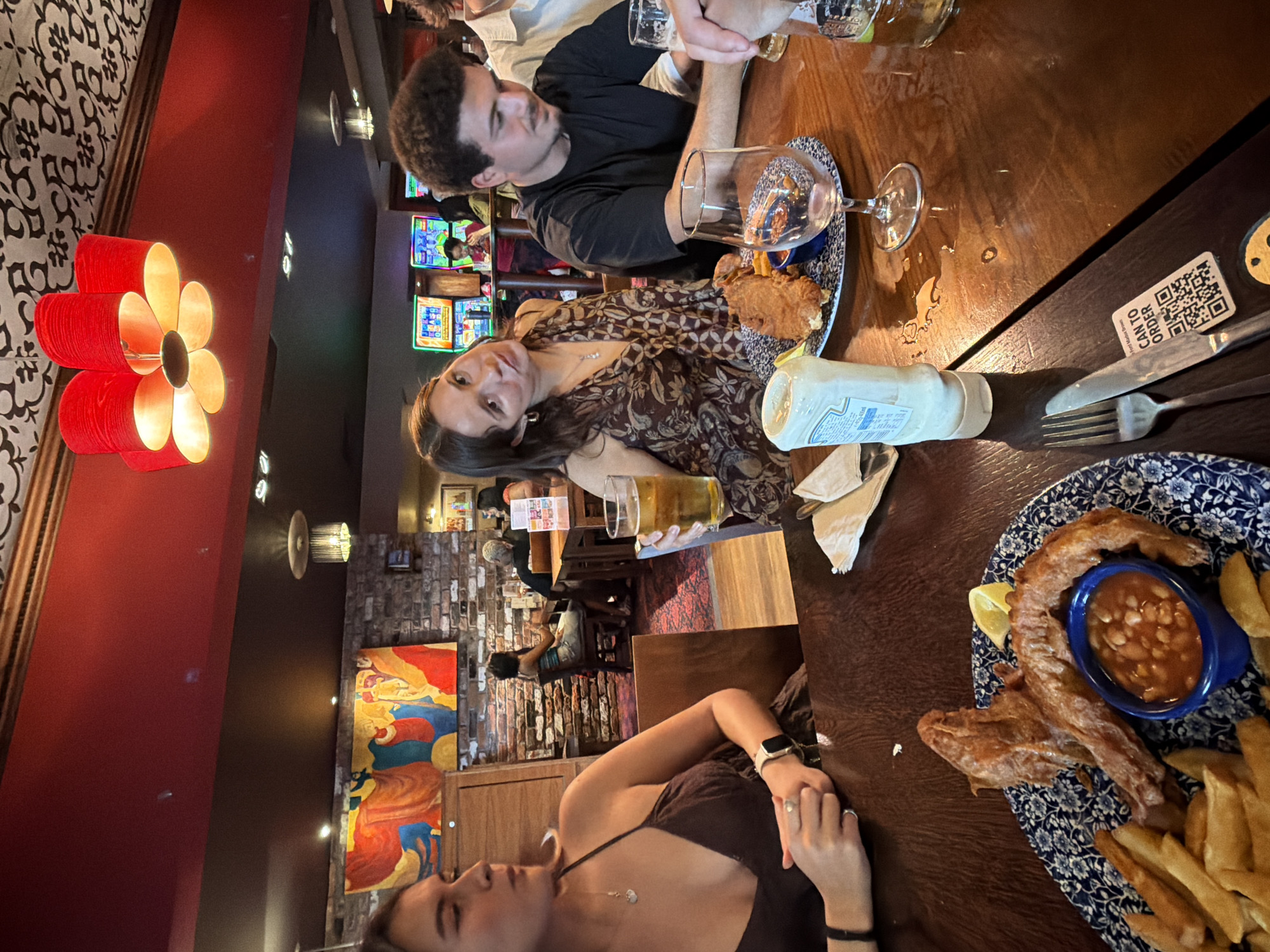 The scene is an indoor shot, likely taken in a restaurant or pub with a distinctive red and black interior theme, featuring decorative floral lighting fixtures. A group of people is seated around a wooden table laden with food and drinks. In the foreground, a woman with dark hair is wearing a black top and a wristwatch. She is situated near a plate of fish and chips, accompanied by a side of beans. A bottle of mayonnaise is also present on the table. Another woman with brown hair, wearing a patterned top, is seated across from her, posing for the camera. A man with curly hair, wearing a black shirt, is next to her, engaged in conversation or observation. A glass of beer is being held by someone at the table, indicating social interaction. In the background, a brick wall adorned with a colorful painting is visible, along with other patrons seated at tables and slot machines lining the wall, adding to the ambient activity. The presence of a QR code on the table suggests a method for ordering or accessing information. The environment suggests a casual dining experience, possibly in the city of Unknown, Unknown.