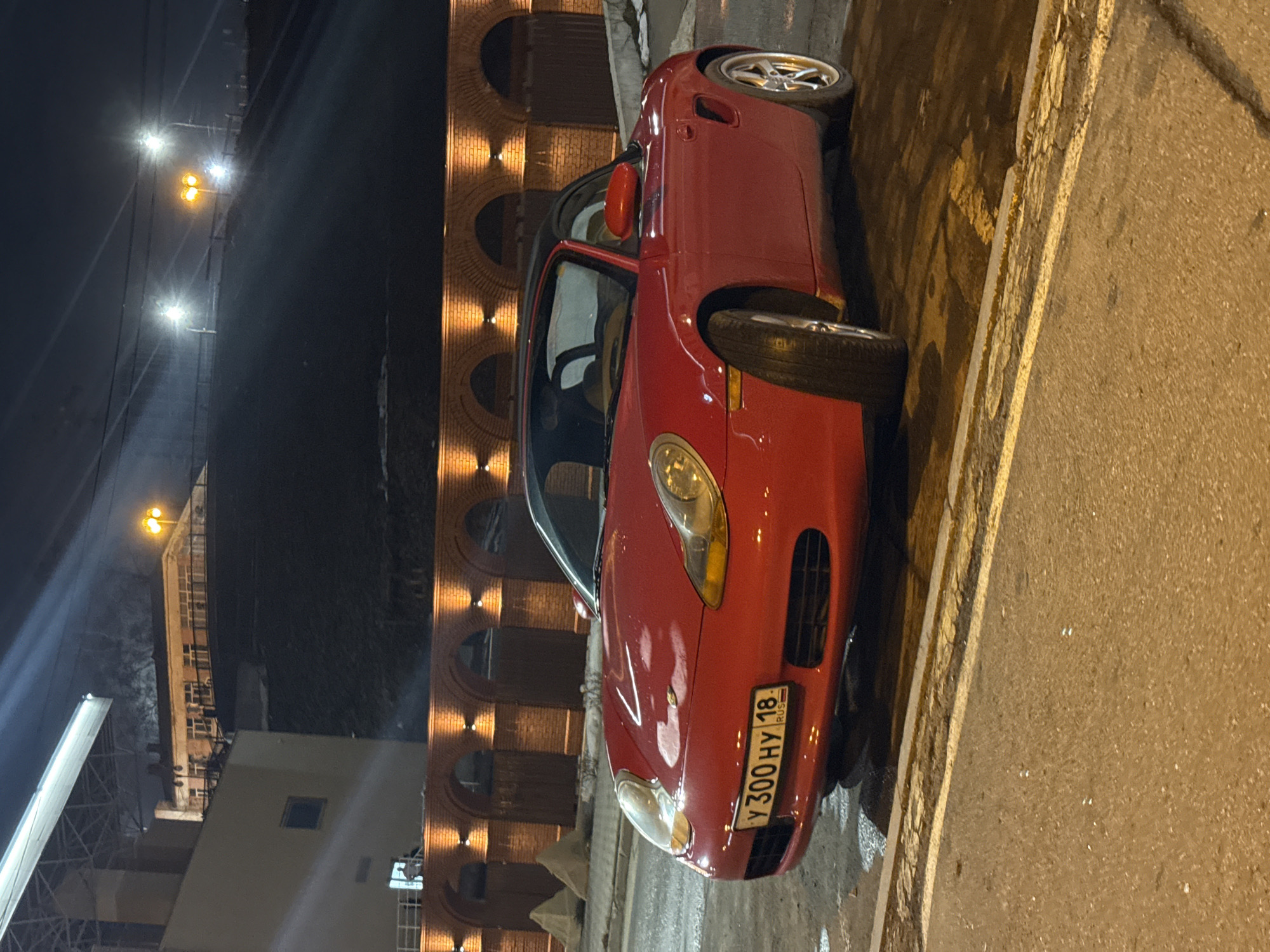 A red convertible car, likely a Porsche, is parked on a street adjacent to a building with arched windows and decorative lighting. The license plate on the car reads "У 300 НУ 18 RUS", indicating the car is registered in the Udmurt Republic of Russia, where Izhevsk is located. The car is situated on a paved surface, possibly a road or parking area, near a curb. Street lights illuminate the area. The location is Izhevsk, Russia.