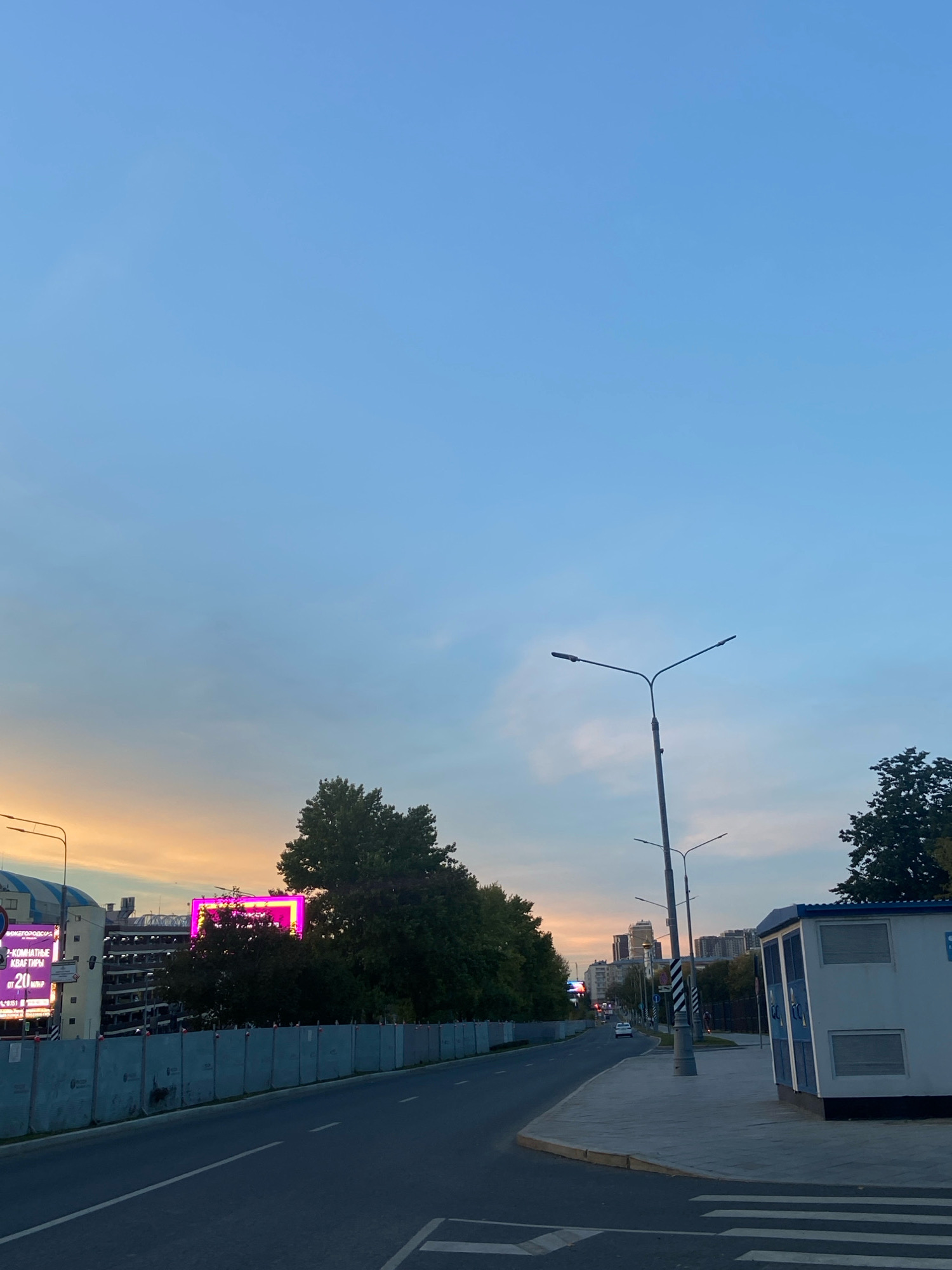 The image depicts an urban scene in Moscow, Russia, likely at dusk given the soft, warm glow on the horizon and the clear, pale blue sky. A wide asphalt road with lane markings curves into the distance. To the left, a construction fence encloses an area with what appears to be a multi-level parking garage and large billboards. One billboard features bright pink neon lights outlining a square and displays text in Russian. Further down the road on the left, trees are visible. On the right side of the road, a paved sidewalk runs alongside, bordered by a white and blue utility structure with ventilation grates. Tall streetlights with multiple lamps are spaced along the road. In the background, city buildings of varying heights are silhouetted against the sky. A few cars are visible on the road. The overall impression is one of a street scene in a developed urban area.