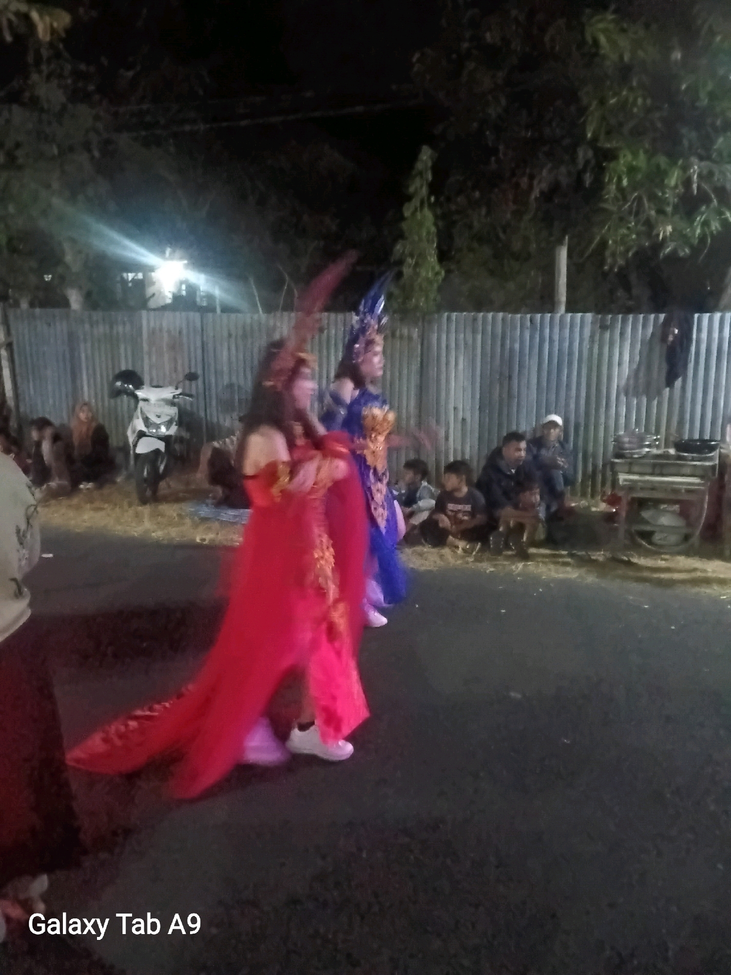 The image depicts an outdoor scene at night, possibly in Wonoanti, Indonesia, featuring a procession or performance. Two individuals in elaborate costumes are the primary subjects. The person on the left wears a long, flowing red dress with gold accents, a feathered headpiece, and white shoes. The person to their right is dressed in a blue costume with a similar feathered headpiece and is partially obscured. They appear to be walking on a dark, paved surface, which could be a road or pathway.
In the background, a corrugated metal fence runs horizontally across the image, separating the performance area from a slightly elevated, grassy or dirt area. Several people are seated or kneeling behind the fence, observing the event. Among them is a white motorcycle parked to the left. Further to the right, a small group of men are seated, and a vendor's cart with cooking equipment is visible. The environment suggests a community event or celebration taking place in a semi-urban or rural setting, with artificial lighting creating shadows and highlights. The overall atmosphere is festive, indicated by the costumes and the gathering of onlookers.