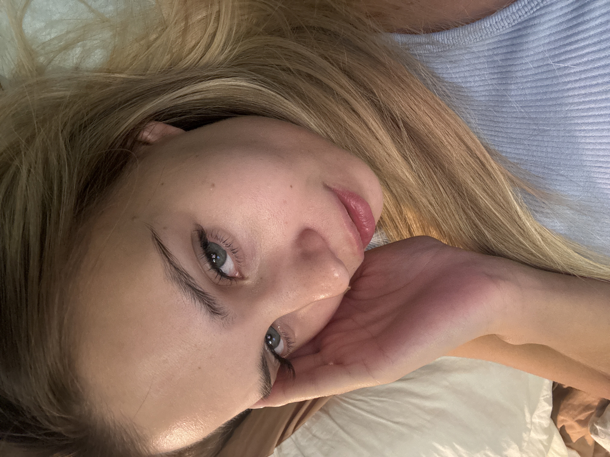 The media file presents a close-up, top-down view of a young woman. She has long, flowing blonde hair that frames her face and spreads out around her. Her light blue-green eyes look directly at the viewer, with distinct eyelashes and defined eyebrows. Her fair skin shows a few subtle moles on her forehead and cheek, and her lips are a natural pink.
The woman is lying down, likely on a bed, as suggested by the white pillow or sheet visible beneath her and a hint of brown fabric under her arm. She is wearing a light blue, ribbed tank top or camisole. Her right hand is gently positioned beneath her jawline, appearing to support her head in a relaxed pose.
The lighting is soft and natural, highlighting her features and creating a subtle glow on her skin. Her expression is calm and serene, conveying a sense of repose or contemplation. The setting appears to be indoors, possibly a bedroom, but there are no specific visual cues within the image itself to indicate its location as Chaikovsky, Russia. No text is visible in the image.