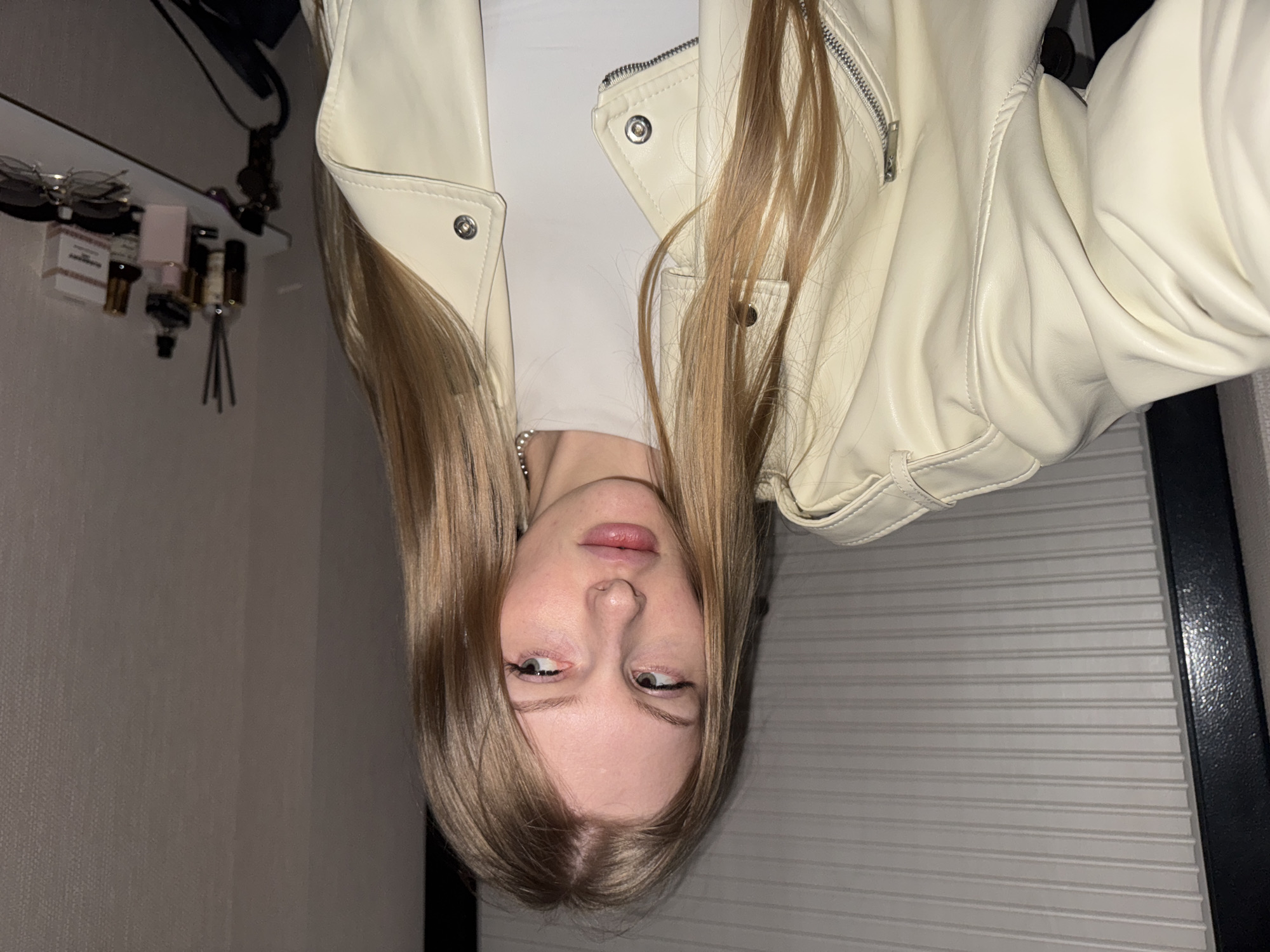 An inverted portrait features a young woman with long, straight, light brown hair, appearing to be in her late teens or early twenties. She has fair skin, full lips, and her eyes glance slightly upwards and to the sides, conveying a somewhat neutral or contemplative expression. She wears a light cream or off-white leather/faux leather jacket with metallic zippers and snap buttons, over a white top. A delicate silver chain is visible around her neck.
The setting is an indoor room, likely a bedroom or personal space. In the background, on what would be the top-left if the image were rotated, a narrow shelf holds several items, including sunglasses and various bottles, some appearing to be perfumes or essential oils with diffuser sticks. One small box on the shelf partially displays the word "CHANEL." A black strap or bag hangs from above the shelf. The walls are a plain, light grey or beige. To the right of the woman, a vertically ridged panel, possibly a radiator cover or blind, abuts a dark, vertical frame.
The image captures a close-up pose, suggesting a selfie or a directed portrait. The lighting appears to be artificial, indicative of an indoor setting, possibly during the evening or night. The overall impression is an intimate, casually posed shot, taken in Perm, Russia.