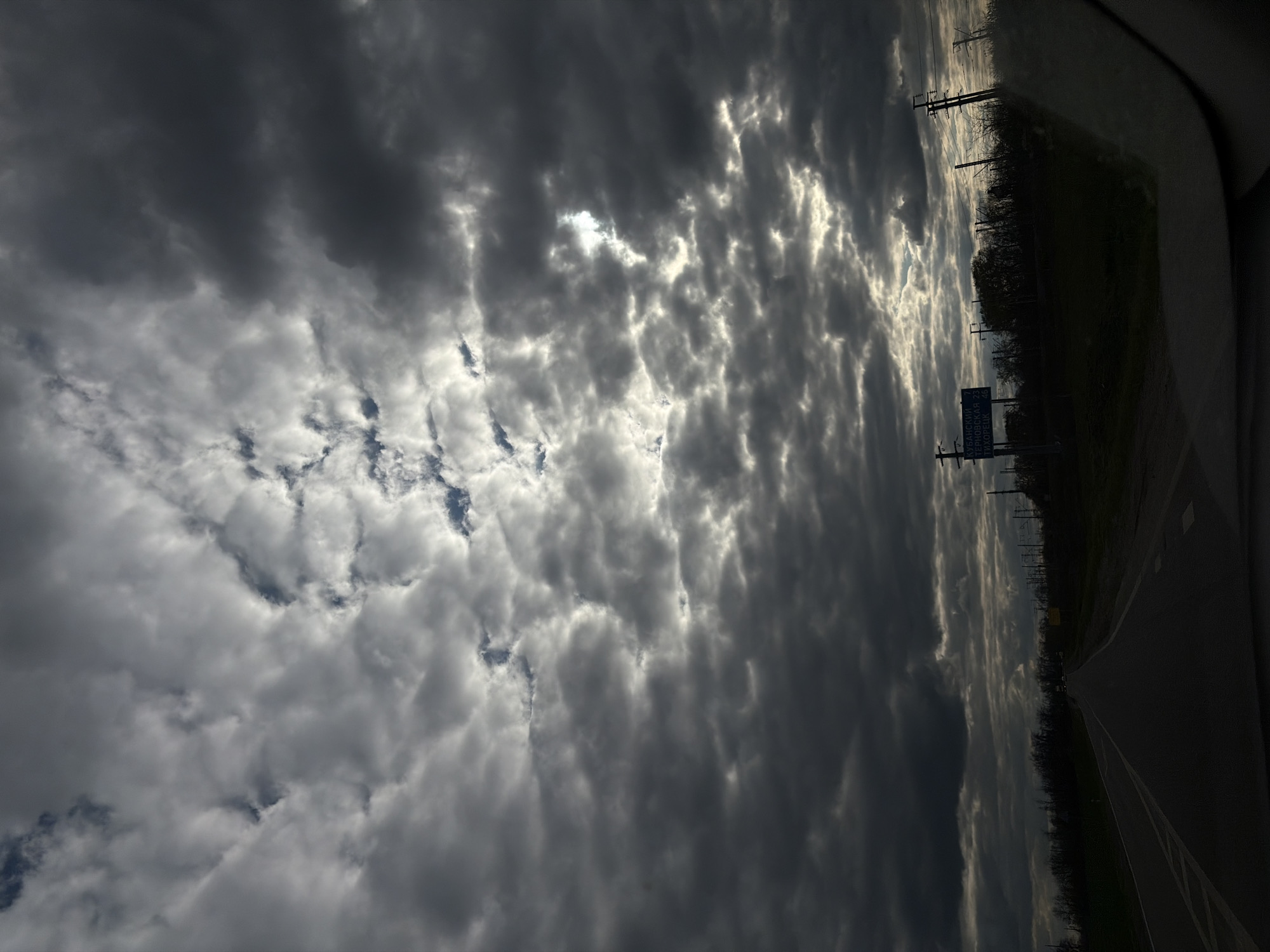 The image captures an overcast sky above a rural road, likely taken from inside a vehicle. The scene is oriented vertically, but when viewed horizontally (rotated 90 degrees counter-clockwise), it shows a dramatic sky filling the majority of the frame, with a dark roadside and a road extending into the distance on the right.
The sky is dominated by dense, grey clouds, with patches of bright light breaking through, suggesting the sun is hidden but casting strong illumination on certain cloud formations. Some areas of blue sky are visible through gaps in the clouds.
Along the right side, a dark strip of land represents the roadside, featuring silhouetted trees, bushes, and multiple utility poles with power lines. A paved road, marked with faded white lines, curves slightly into the distance.
A blue road sign stands prominently on the right side of the road, displaying white Cyrillic text. The text reads:
- КУБАНСКИЙ 7
- ТЕРНОВСКАЯ 25
- ГУЛЬКЕВИЧИ 36
These are names of settlements (Kubansky, Ternovskaya, Gulkevichi) followed by their respective distances in kilometers, indicating the location is in Russia, consistent with the Novopokrovskaya area context.
The time of day appears to be either late afternoon or early morning, characterized by the dramatic interplay of light and shadow created by the heavy cloud cover. No people are visible in the image. The overall impression is one of travel under a dynamic, brooding sky.