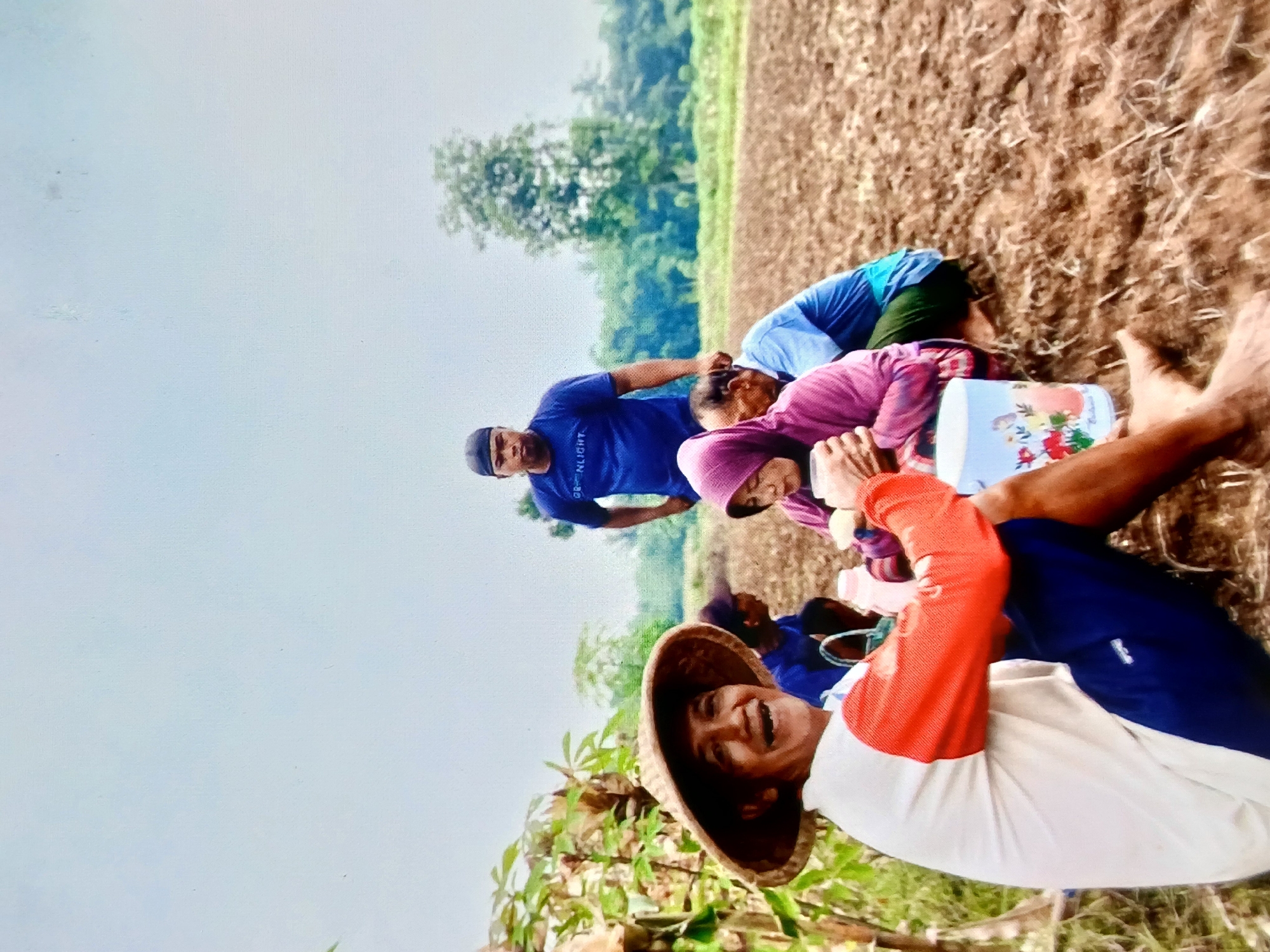 In Setia Bakti, Indonesia, a group of people are gathered in a tilled field under an overcast sky. In the foreground, a man wearing a conical straw hat and a white shirt with orange sleeves, along with dark blue shorts, is seated and smiling broadly. Beside him, a light blue cup with floral designs sits on the ground. Behind him, partially obscured, are several other individuals. A man in a blue t-shirt with the word "NLIGHT" visible stands in the midground. To his right, a woman in a pink headscarf and jacket is seated, holding a white object. Another person in a blue shirt and green pants is also seated in the midground. The environment consists of dry, brown soil, with some vegetation and trees visible in the background.