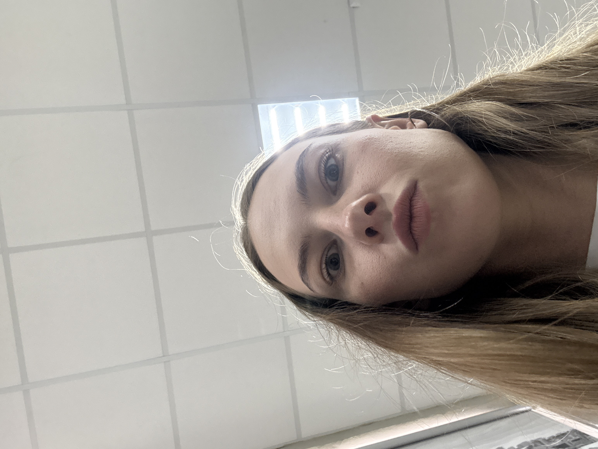 A low-angle, close-up shot shows the face of a young woman with light brown hair. She has wide eyes and her lips are pursed. The background is a white tiled ceiling with fluorescent lighting. The image appears to have been taken indoors. There is no indication of the city of Perm, Russia, or any other specific location.