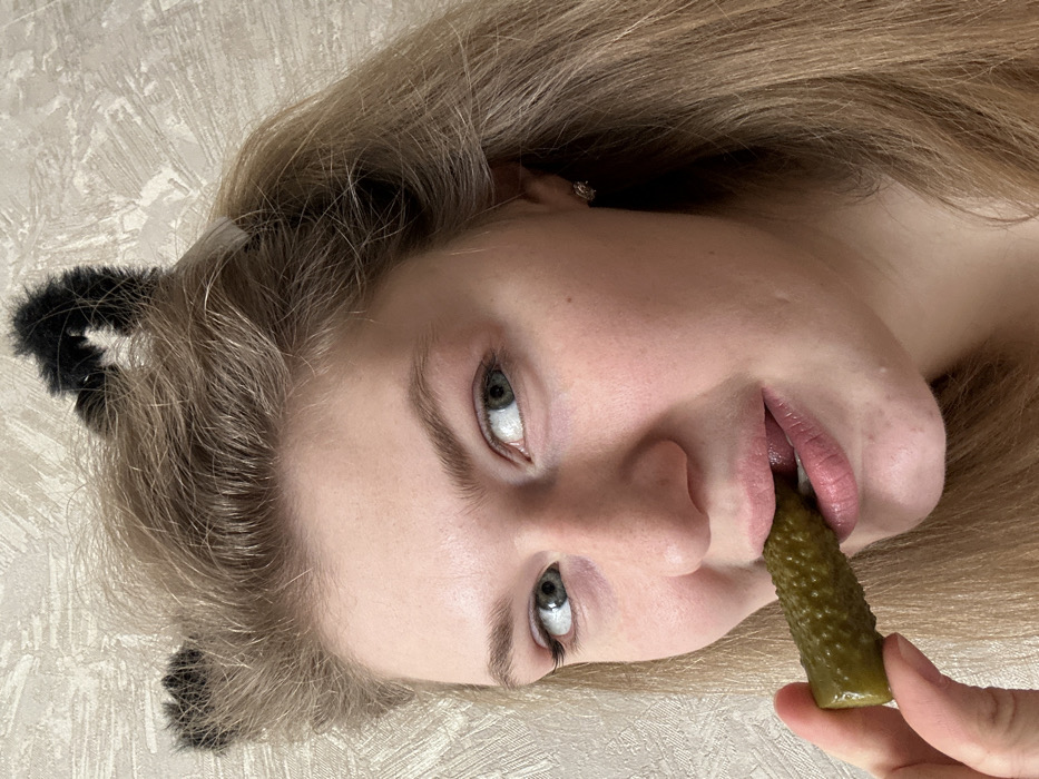 A young adult female individual with light-colored hair and fair skin is depicted in a close-up, chest-up indoor shot. She wears a black furry headband styled as cat ears and small stud earrings. Her light-colored eyes are directed towards the viewer. The subject holds a single green gherkin between her left thumb and index finger, with the gherkin partially inserted into her open mouth. Her tongue is visible against the gherkin. The background consists of a light-colored, subtly textured wall. The visual information available in the image does not provide any specific identifiers to confirm the location as Nizhnekamsk, Russia.