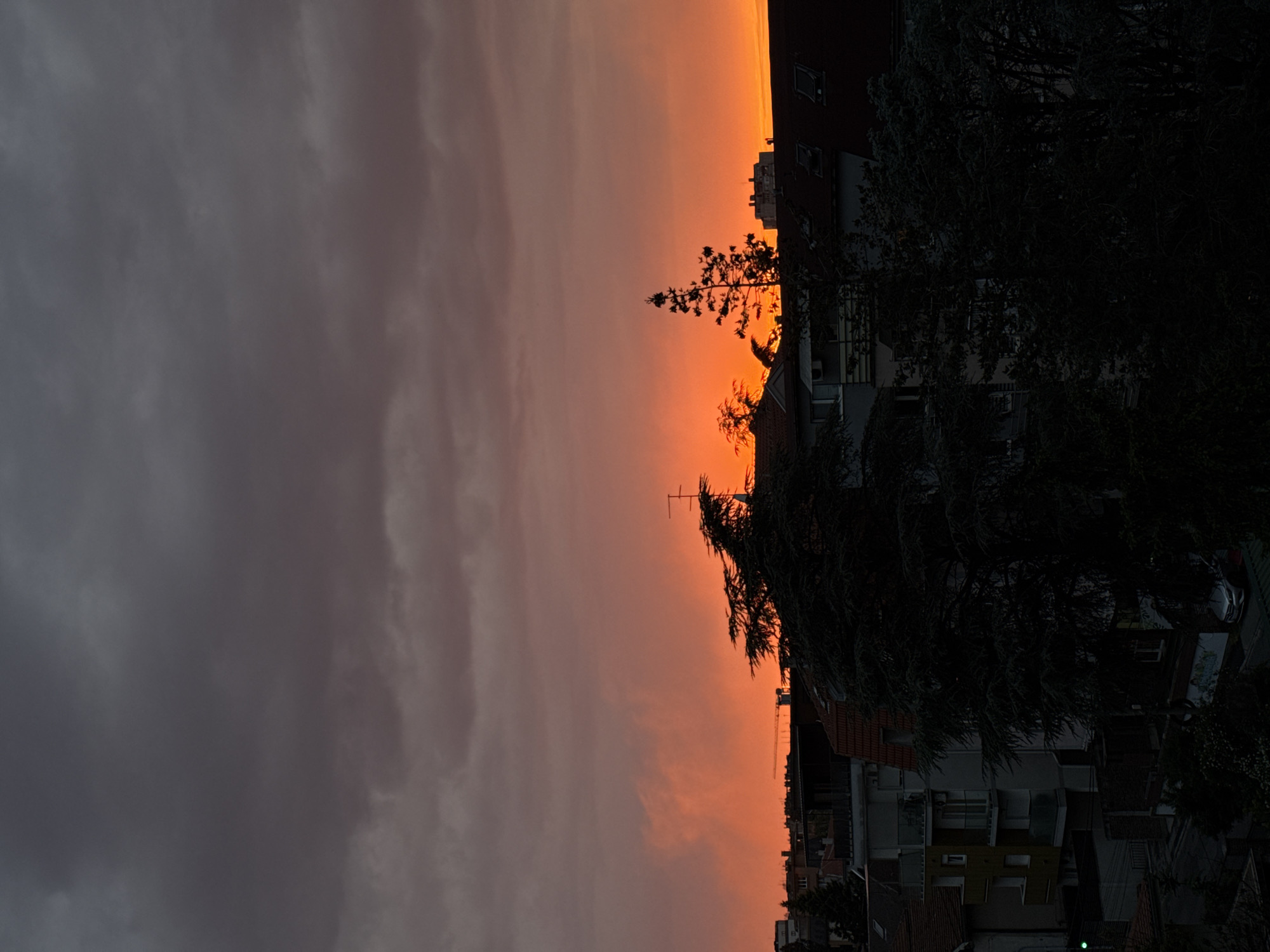 The image depicts a sunset in Vozdovac Urban Municipality, Serbia. The sky transitions from dark clouds on the left to a vibrant orange hue near the horizon. Buildings and trees are silhouetted against the bright sky. An antenna is visible atop one of the buildings. The scene appears to be a residential area.