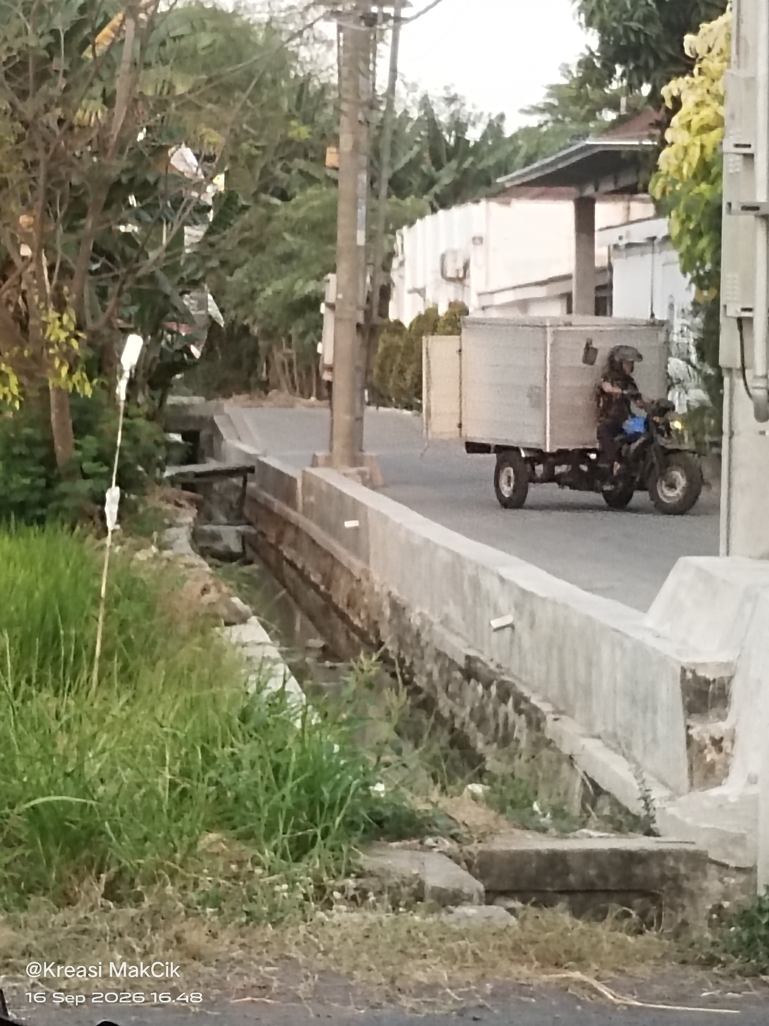 A person wearing a helmet is riding a three-wheeled motorcycle with a large cargo box attached to the back. The motorcycle is on a paved road in what appears to be a rural or suburban area. To the left of the road, there is a drainage ditch filled with water and overgrown with grass and vegetation. Further to the left, there are trees and foliage. To the right of the road, there is a concrete barrier or wall. In the background, there are buildings and more trees. The sky is overcast. The image is dated 16 Sep 2026 16.48. The location is identified as Sambungrejo, Indonesia.