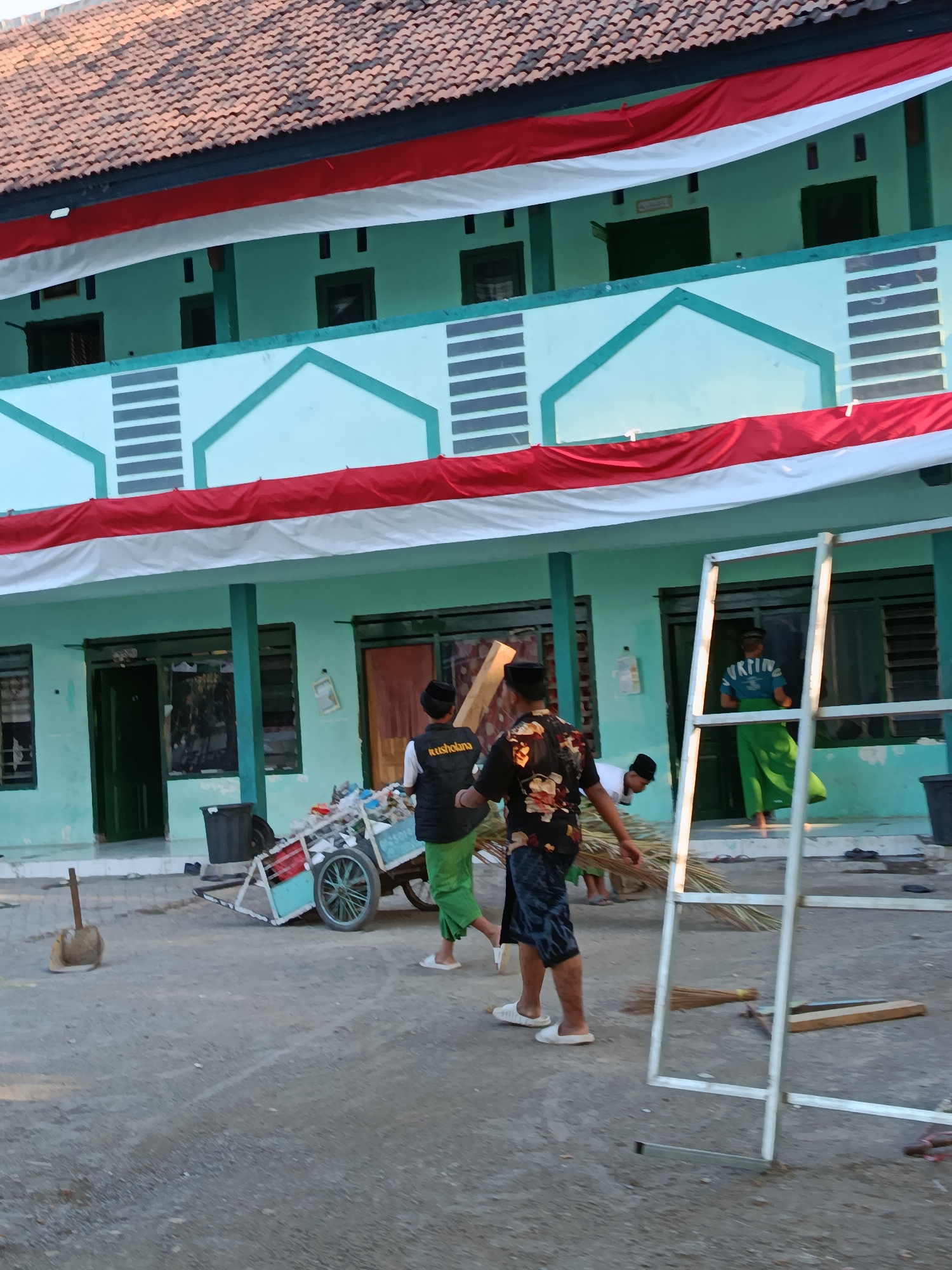 The image depicts an outdoor scene at a building adorned with Indonesian flags, likely in Ramban Wetan, Indonesia. In the foreground, a person is pushing a wheelbarrow filled with what appears to be trash and debris. To the left of the wheelbarrow, a broom leans against a short shovel. In the background, several individuals, predominantly young males wearing traditional Islamic caps (peci) and loose-fitting attire, are engaged in clearing the area. One person is seen sweeping with a broom. The building itself is painted a light green and features decorative white and gray elements. Red and white fabric banners are draped across the building's facade, further emphasizing the national theme. The ground is a rough, dusty surface, indicative of a courtyard or open area.