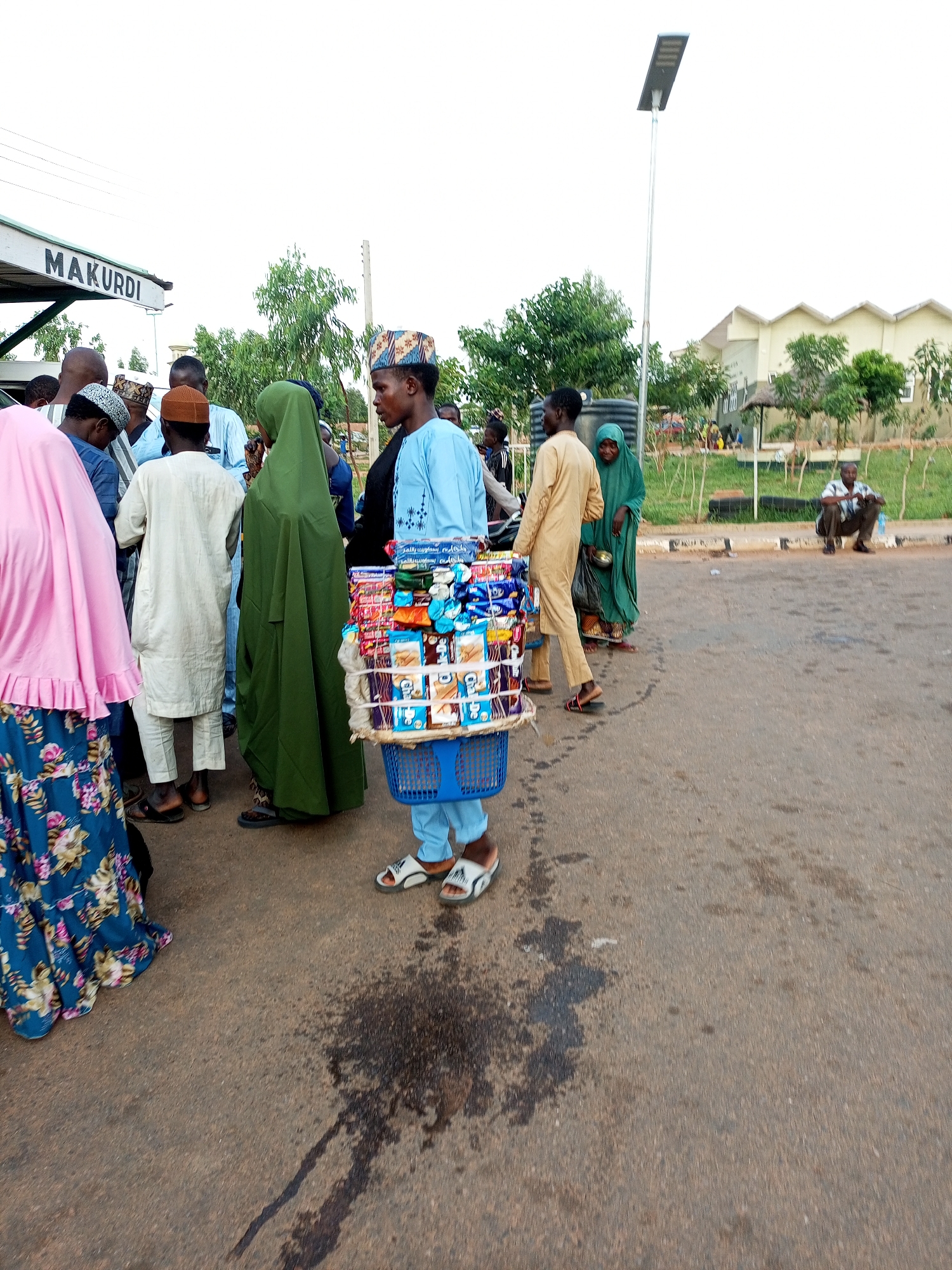 The image depicts a street scene, likely in Gombe, Nigeria, with several individuals present. In the foreground, a young person wearing a light blue tunic and pants is standing, holding a blue basket filled with various packaged snacks and drinks. This person is likely a vendor. To the left, a person in a pink hijab and a floral skirt is visible. Further back, a group of people are gathered, some wearing traditional Islamic attire, including a person in a long green hijab. The background shows a street with a white sign bearing the word "MAKURDI" in black capital letters, suggesting a location or destination. A solar-powered street lamp stands tall on the right side of the image, and a building with a distinctive roofline is visible in the distance, surrounded by trees and greenery. The ground appears to be asphalt, with some dark stains indicating spillage. The overall atmosphere suggests a public space, possibly a market area or a transportation hub.