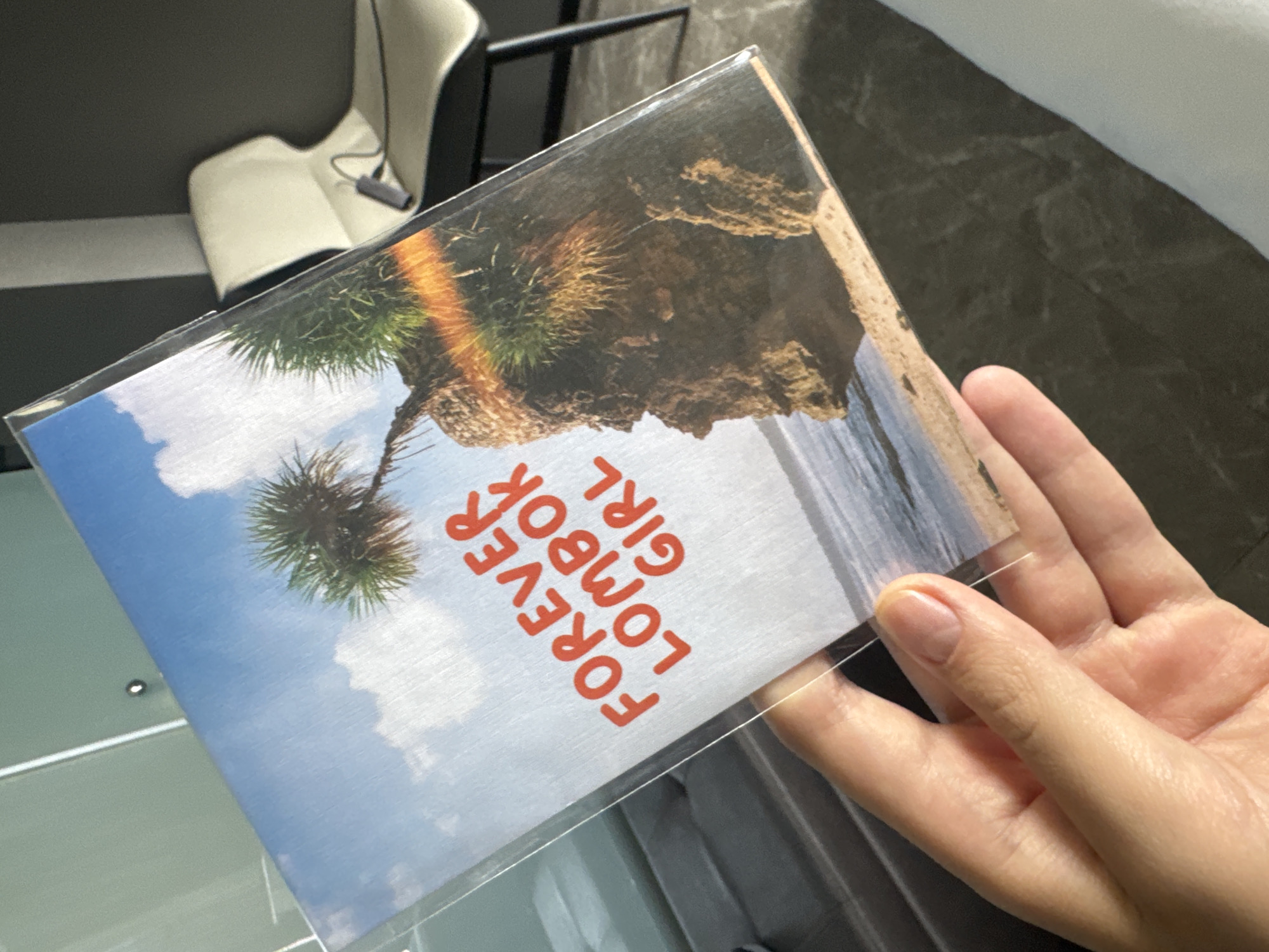 A hand holds a postcard in a clear plastic sleeve. The postcard features an image of a tropical landscape with a rocky cliff, palm trees, and the ocean. The text "FOREVER LOMBOK GIRL" is printed in red, rotated 90 degrees, and stacked vertically. The hand is holding the postcard against a background that includes a white chair and a gray wall. The location depicted on the postcard is likely a coastal scene in Lombok, Indonesia, which is near the city of North Kuta.
