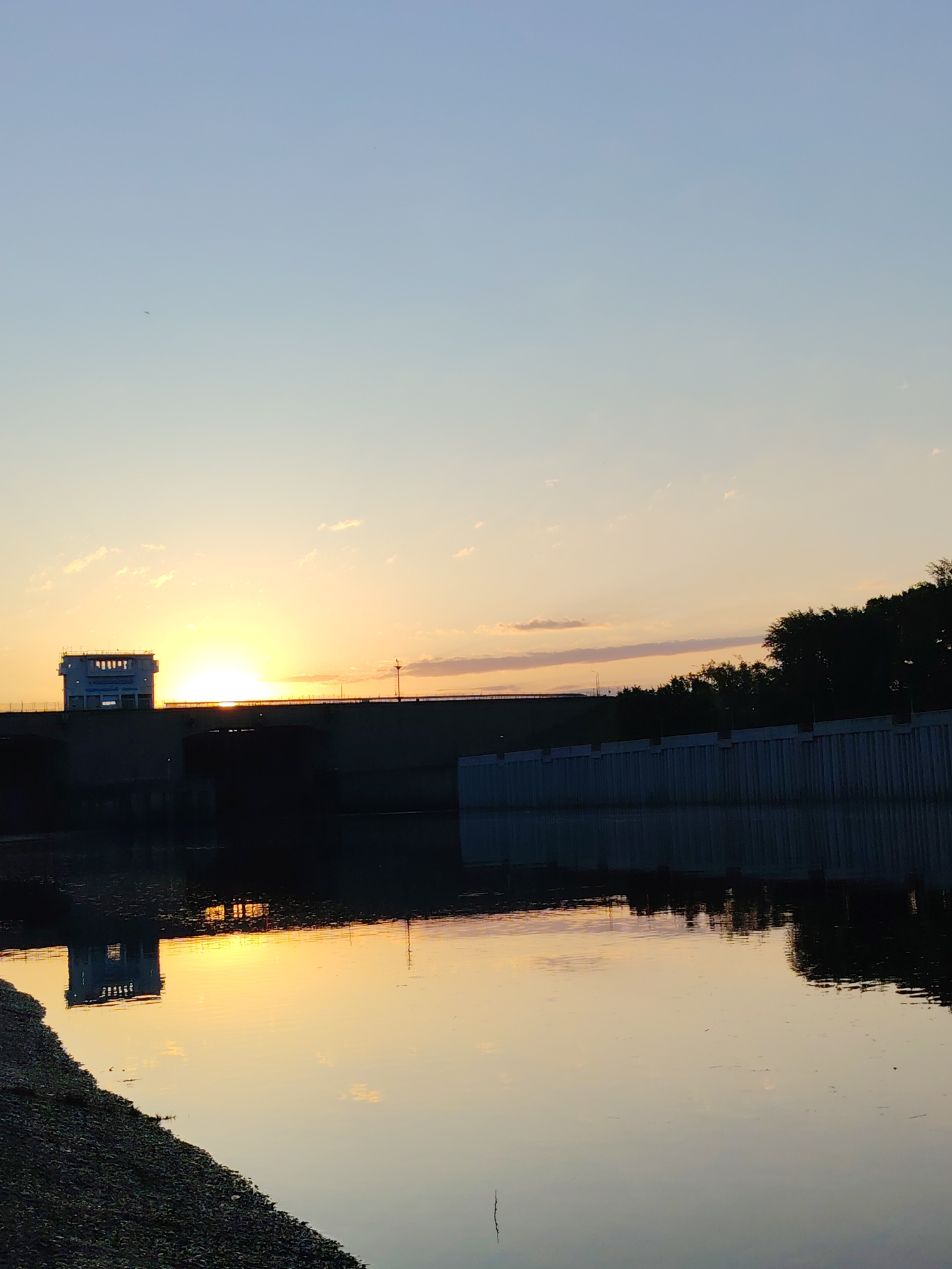 The image shows a sunset over the Kama River near the city of Chaikovsky, Russia. The sun is positioned just above the bridge, casting a bright light and creating a reflection on the water. A building sits on the left side of the bridge. The right side of the bridge is bordered by trees. The water is mostly still with the bridge, building, trees, and sky reflected on it. There is some gravelly shoreline in the lower left.