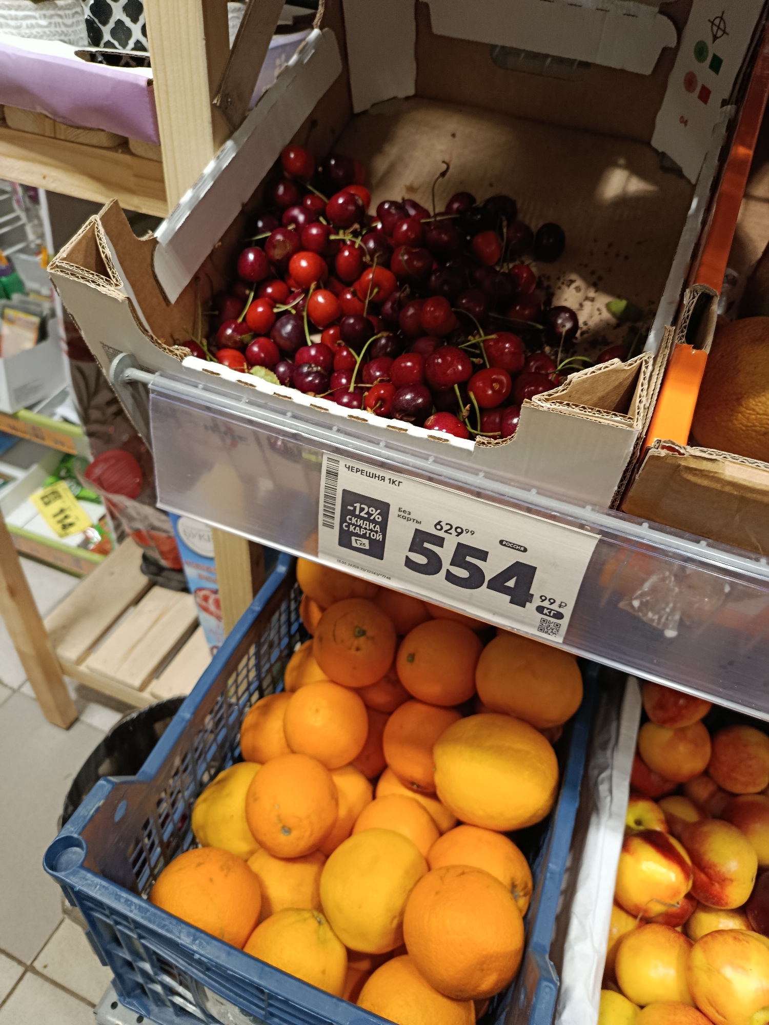 A close-up, eye-level shot captures a display of fresh produce at a market, possibly in Verеya, Russia. In the foreground, a blue plastic crate is filled with bright orange fruits, likely oranges, with some yellow fruits, possibly lemons or grapefruits, interspersed. A white price tag is visible, displaying the text "ЧЕРЕШНЯ 1КГ" (Cherries 1kg) and indicating a price of "554". Above this, a cardboard box is overflowing with fresh, dark red cherries, their stems attached. The cherries are piled high, suggesting a generous portion. In the background, other produce and store shelves are partially visible, indicating a retail environment. The lighting appears to be from overhead fixtures, casting a soft glow on the fruits.