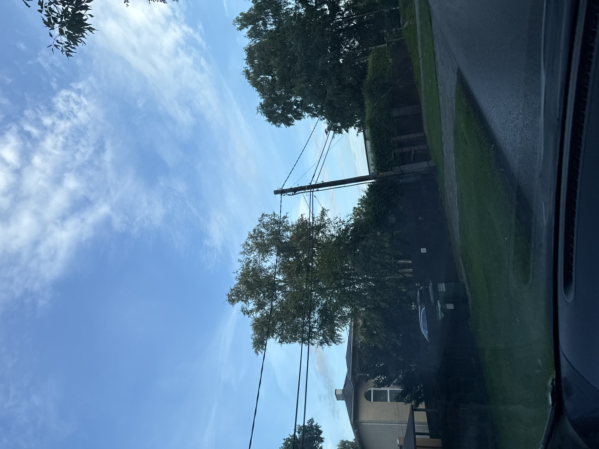 The image depicts a street view in Tallinn, Estonia, under a partly cloudy blue sky. In the foreground, the dashboard of a vehicle is visible, suggesting the photo was taken from inside a car. The middle ground shows a street with a paved surface, bordered by a grassy area and a neatly trimmed hedge. Beyond the hedge, a modern house with a light-colored facade and a distinctive arched window is partially visible, surrounded by trees. A dark-colored car is parked in front of the house, near a green dumpster. A utility pole with multiple wires runs across the scene. The overall impression is one of a quiet, residential suburban area.