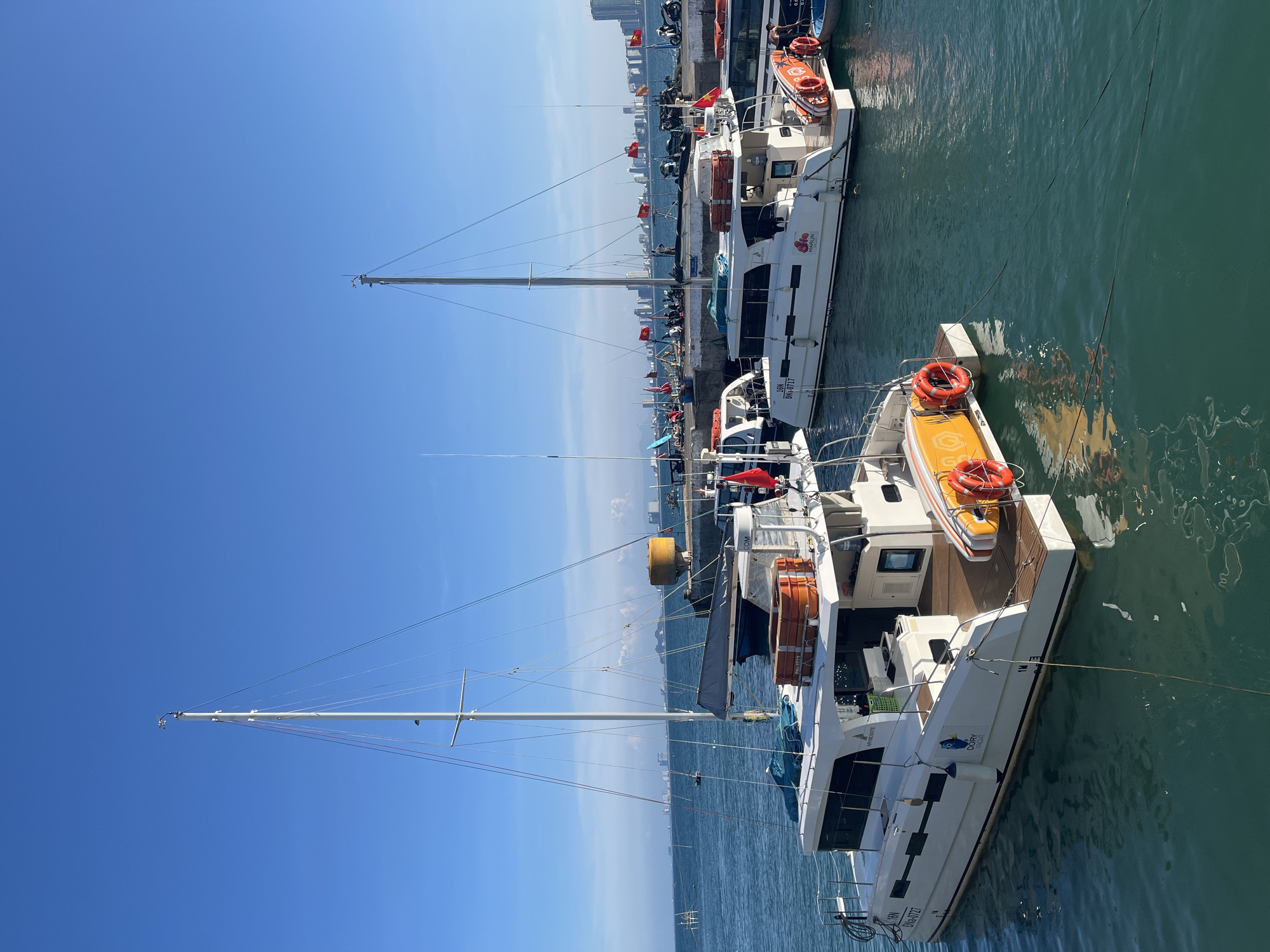 The image shows a marina in Đà Nẵng, Vietnam, filled with yachts and boats docked alongside a pier. Clear blue sky is visible above. The water in the marina is green. Buildings are visible in the background. A series of Vietnamese flags line the pier. The boats have white hulls and masts. Some are equipped with orange life preservers and rescue rafts. Mooring lines connect the boats to the pier.