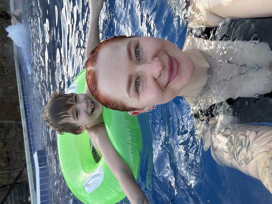 An outdoor swimming pool scene in Rostov-on-Don, Russia, features two individuals. In the foreground, an adult female with wet, reddish-orange hair, light-colored eyes, and visible freckles on her face and shoulders is taking a selfie. She smiles directly at the camera, displaying a septum piercing, multiple earrings in her left ear, and wearing a dark-colored swimsuit. A silver-toned necklace with a small pendant hangs around her neck, and a patterned tattoo is visible on her left arm. Behind her, partially submerged in the water, is a male child, estimated to be 7-10 years old, with wet brown hair and a wide smile revealing a gap between his front teeth. He is supported by a bright green inflatable swimming ring, with his arms extended. A thin dark cord or necklace is around his neck. Both are in clear, rippled blue pool water. The tiled edge of the pool, featuring blue and white patterns, is visible behind the child. In the background, a textured, brownish-grey wall or structure is present, along with a distant water feature generating a splash.