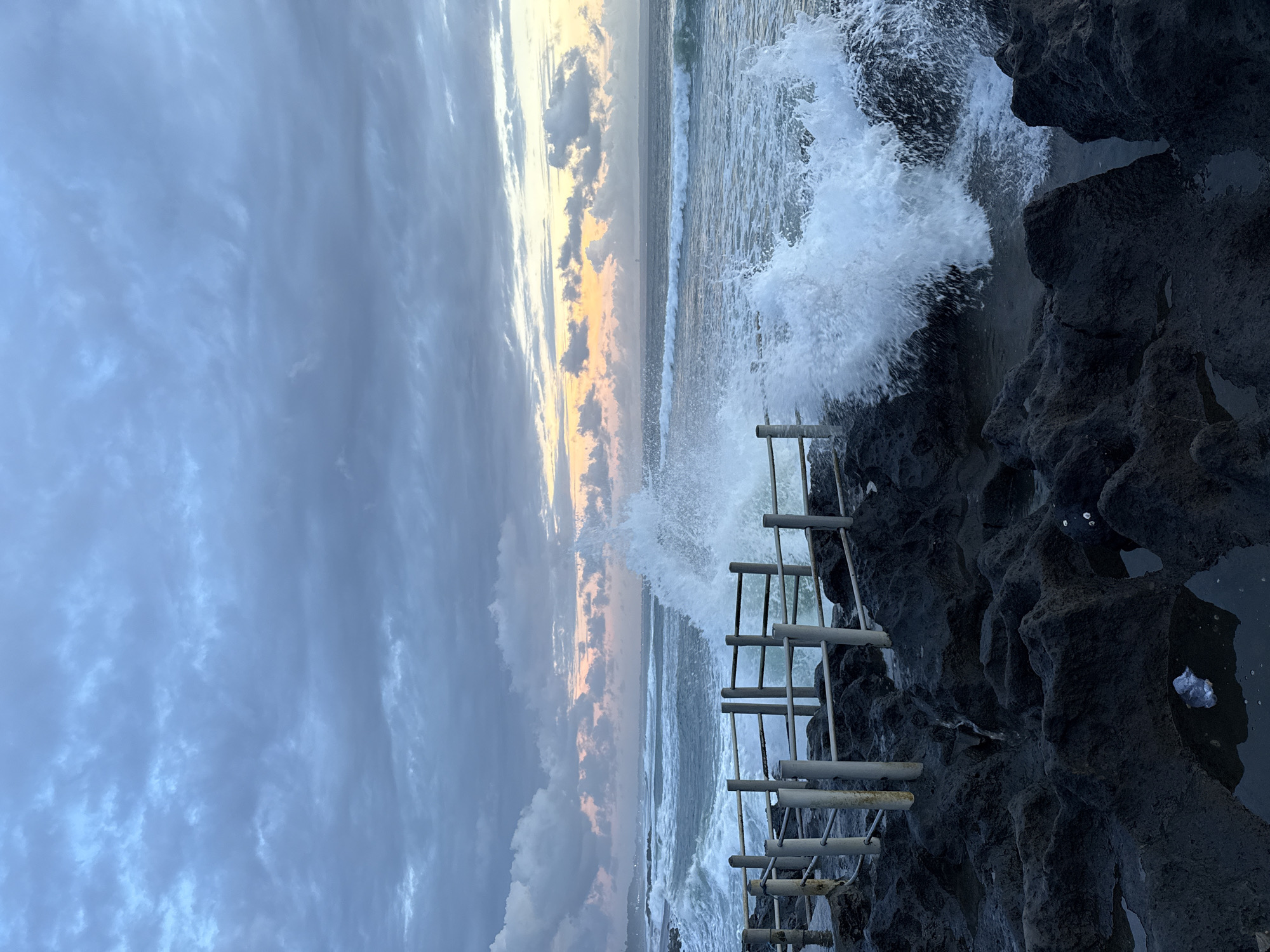 The image shows a seascape in Mengwi, Indonesia. A rough rocky coast is to the right, with an artificial metal barrier structure attached to the rocks. Waves are crashing against the rocks and structure. The sea stretches out to the horizon in the center, where the sun appears to be setting or rising. The sky is covered with grey clouds tinged with orange from the sun.