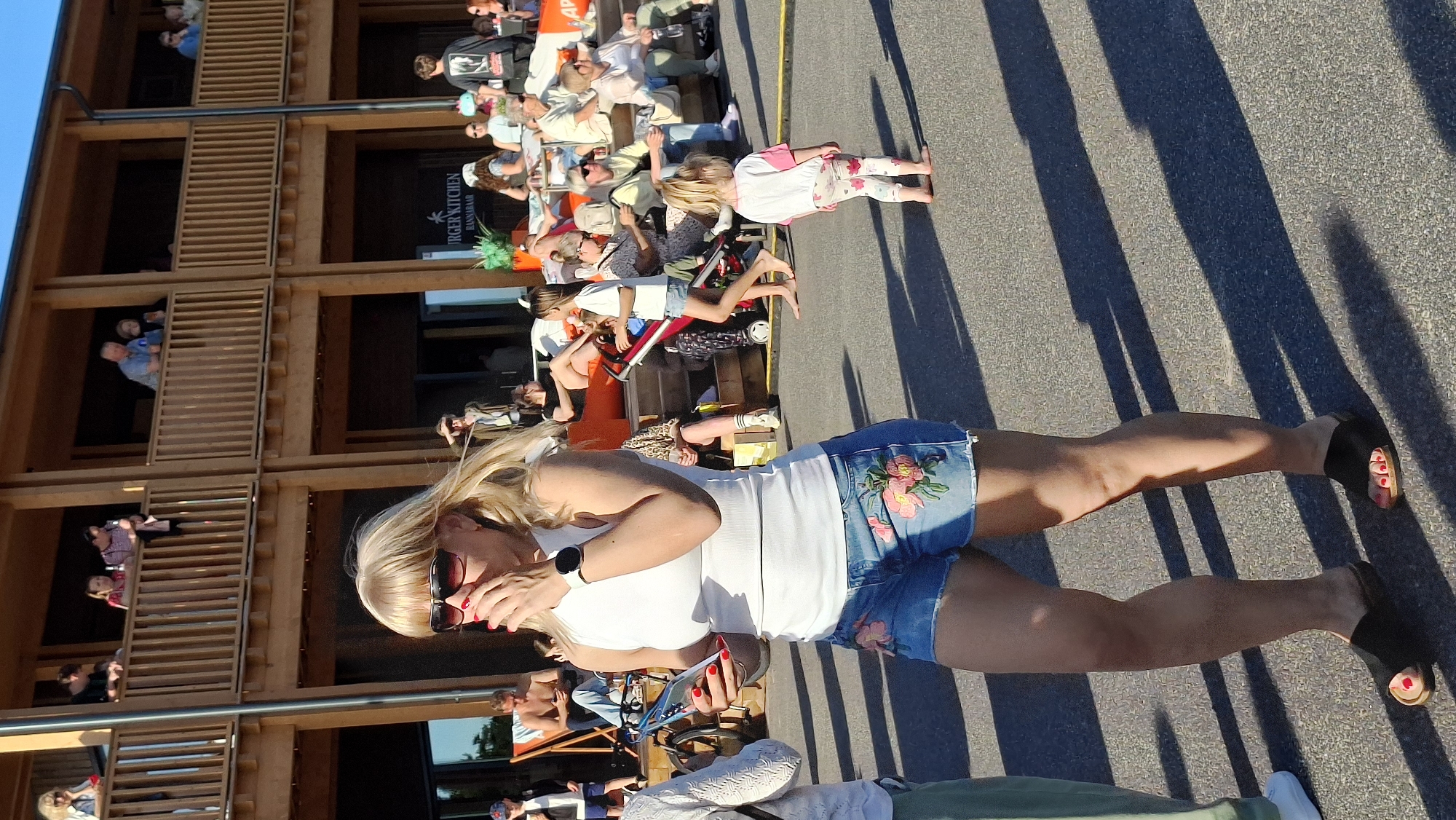 The image depicts an outdoor scene with several people in the foreground and background. In the immediate foreground, a blonde woman wearing sunglasses, a white tank top, and denim shorts with floral embroidery is holding a smartphone and covering her mouth with her hand. She is wearing black slide sandals with red-painted toenails. To her left, another person's legs are visible, wearing a white shoe and green pants.
In the midground, a young girl with blonde hair, wearing a white and pink t-shirt and patterned leggings, stands barefoot facing away from the camera. Behind her, a slightly older girl in a white t-shirt and denim shorts walks with a stroller. Other people are visible sitting and standing on a wooden multi-level structure in the background. The structure has wooden balconies and appears to be part of a building with the words "BURGER KITCHEN HANNA" visible. The ground is paved with asphalt, and long shadows suggest it is a sunny day.
The overall setting appears to be a public outdoor area, possibly a promenade or a gathering space adjacent to a food establishment. Given the context of "BURGER KITCHEN HANNA", it is plausible this scene is located in Tallinn, Estonia, potentially near a waterfront area or a popular urban leisure spot.