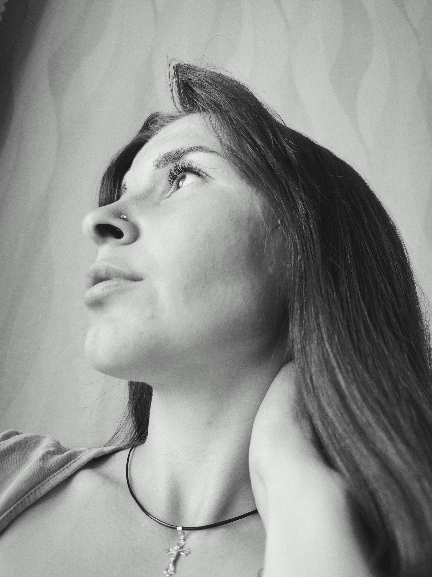A black and white, low-angle portrait features a young woman captured in profile, looking upwards and to the right. The image focuses on her head and upper torso. She has long, dark, straight hair, which is visible flowing down her back and over her right shoulder. A small, shiny stud is visible as a nose piercing in her left nostril. Her prominent eyelashes are clearly visible as she gazes intently upwards. Around her neck, she wears a simple dark cord necklace from which a detailed, silver-colored cross pendant hangs. Her left hand is subtly placed on the back of her neck or upper shoulder. She is wearing a light-colored, possibly fabric, garment visible on her left shoulder. The background is softly blurred, displaying a light tone with subtle vertical or wavy textures, suggesting a curtain or textured wall. The lighting is soft and even, highlighting the contours of her face and the texture of her hair. Her expression appears serene or contemplative, as if lost in thought or observing something beyond the frame. There is no visible text in the image. Location: Фоки, Russia.