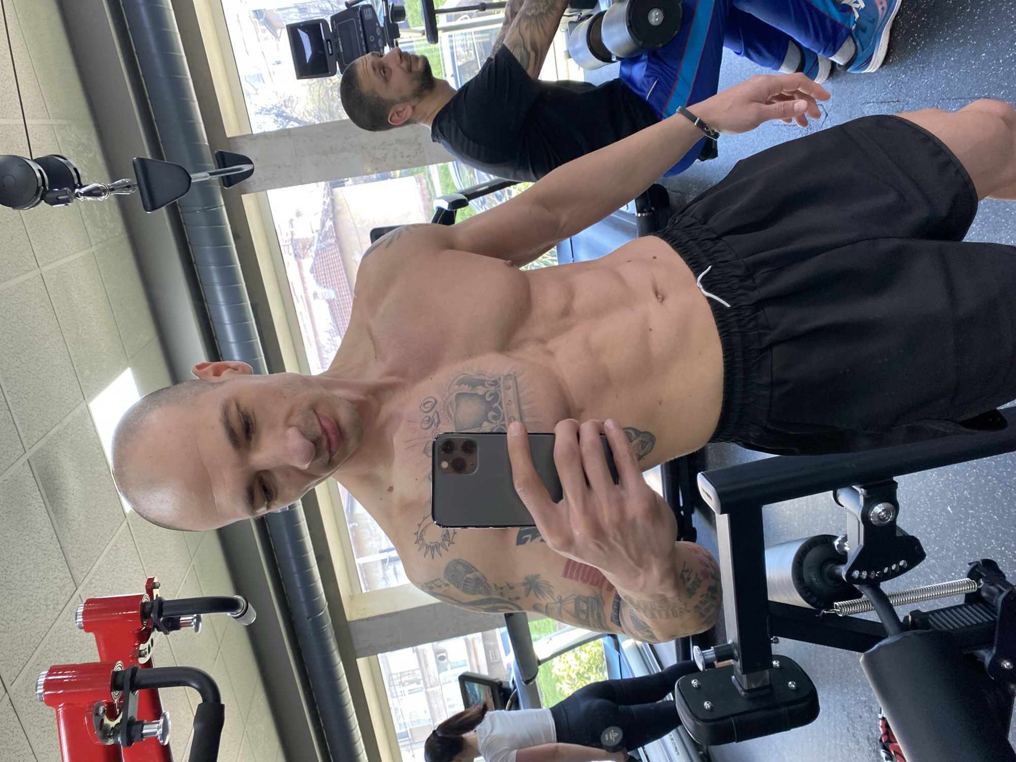 A bald man with tattoos is taking a selfie with a phone in a gym. He is shirtless, wearing black shorts, and has visible abdominal muscles. Another man is lifting weights in the background. A woman is exercising in the background and appears to be on a leg extension machine. The gym has equipment such as weights, cables, and machines. There are windows in the background, showing an urban scene. Based on the man's location data, this scene is in Pleven, Bulgaria.