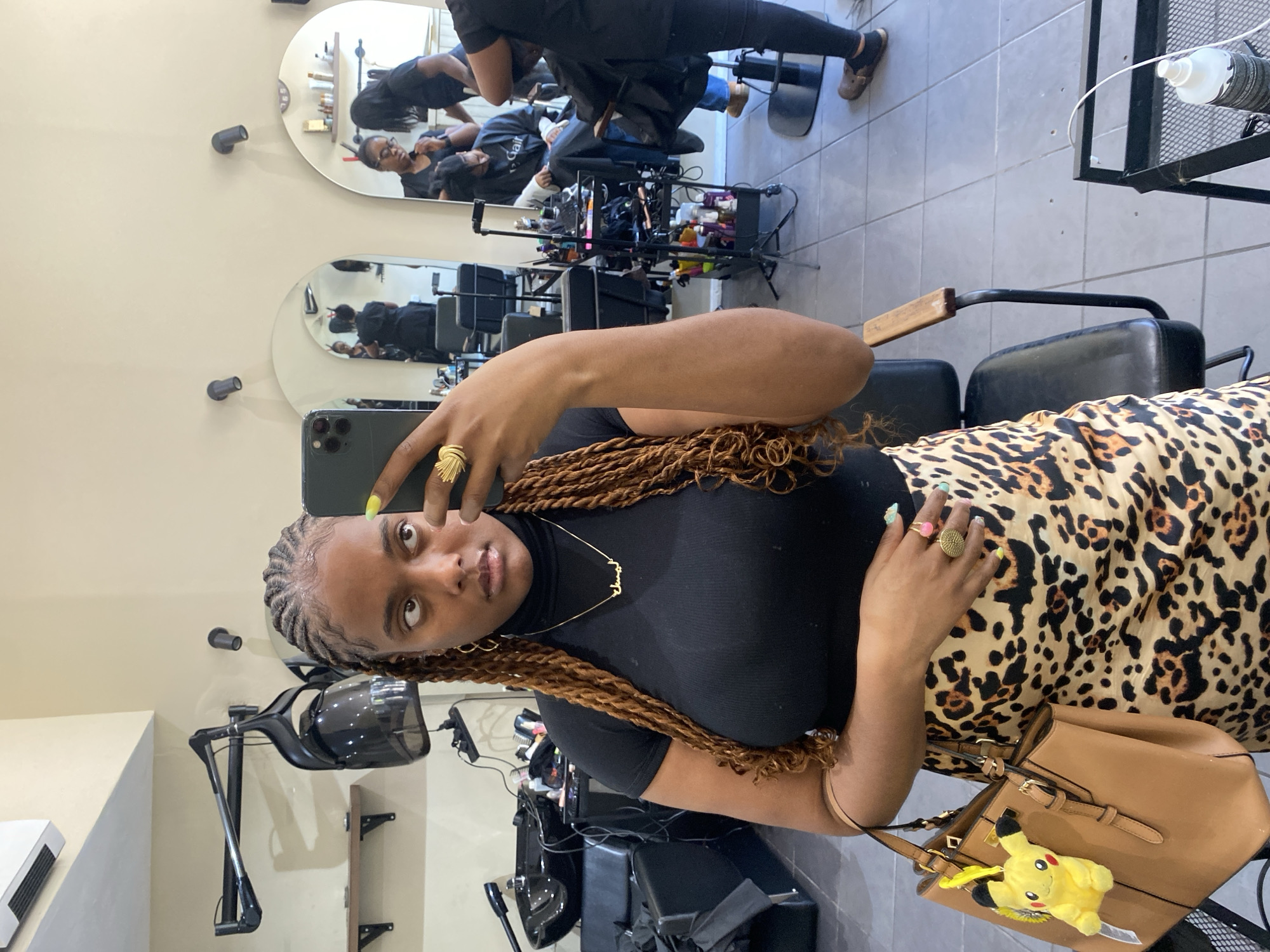 A young Black woman is captured in a selfie taken within a hair salon, with the image originally oriented sideways. She looks directly at the camera, holding a dark-colored smartphone. Her hair is styled in long, reddish-brown twists, with cornrows visible at her hairline. She is dressed in a black turtleneck top and a leopard print skirt, accessorized with a gold-colored necklace (possibly reading "Maria") and multiple rings, including a distinct fan-shaped ring on her right hand. Her nails feature bright polish, with yellow on her right thumb and light blue on her left fingers. A light brown handbag with a yellow Pikachu keychain rests beside her.
The setting is a hair salon, characterized by light beige walls and a light gray tiled floor. Salon equipment such as black styling chairs, a mobile utility cart laden with hair products, and a black stand-alone hair dryer are visible. Two oval mirrors reflect the salon activity; in the upper mirror, a person, likely a stylist, attends to a client wearing a black salon cape, on which the partial text "Gair" is discernible. The lower mirror also shows reflections of other patrons or staff. The ambient lighting suggests it is daytime. The woman appears calm and composed while taking the selfie in what is a typical bustling beauty environment in London.