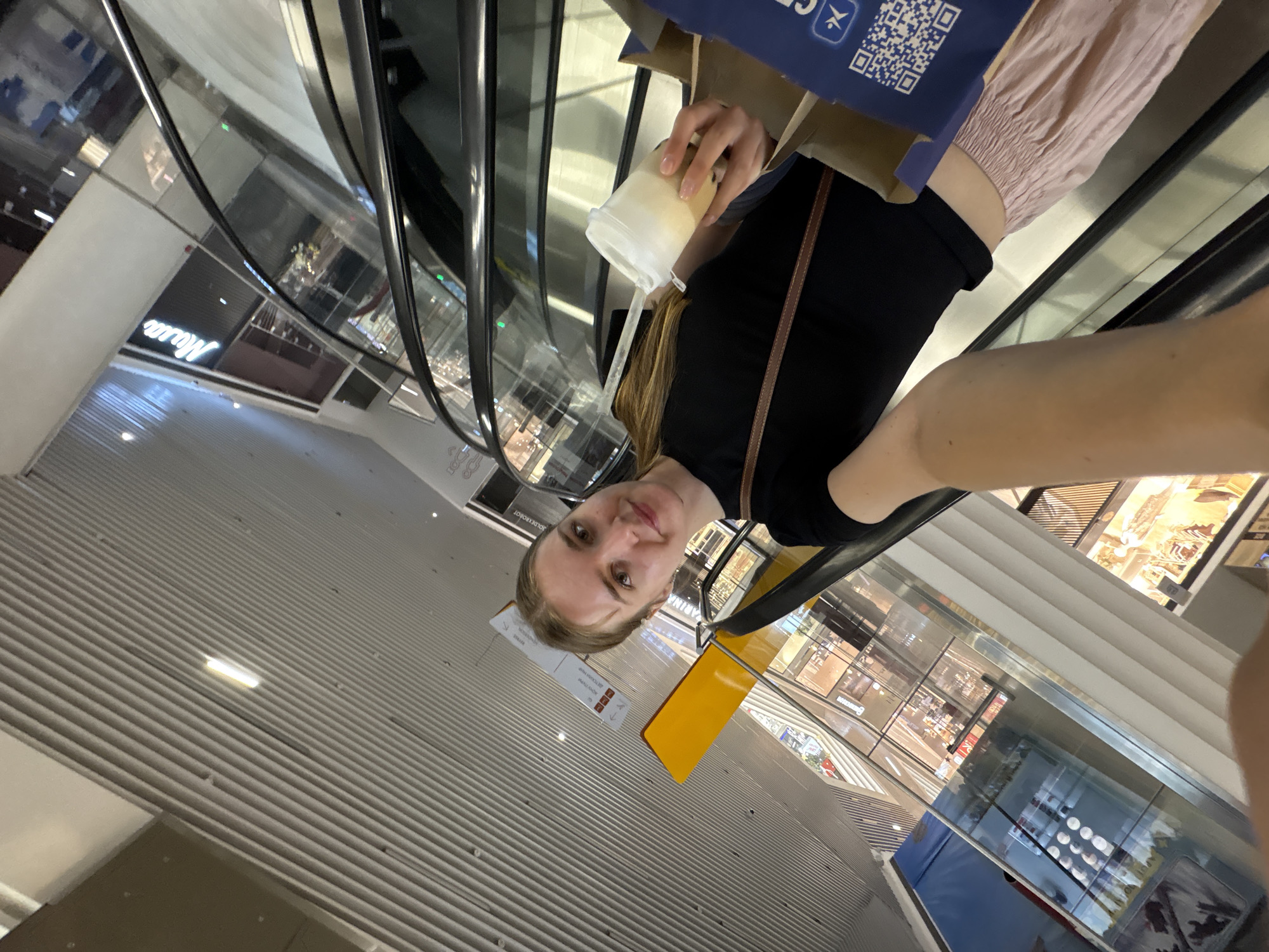 A young Caucasian woman with blonde hair pulled back smiles into a selfie camera while holding a beverage and standing on an escalator. She is wearing a black t-shirt and a light pink skirt. She has a brown strap across her chest from a bag. To her right, a shopping bag is visible with a QR code on it. The background shows the interior of a modern shopping mall, with multiple levels of shops, escalators, and a ribbed ceiling. The signage in the background includes "Masa." The location is identified as the "Masa" shopping mall in Perm, Russia.