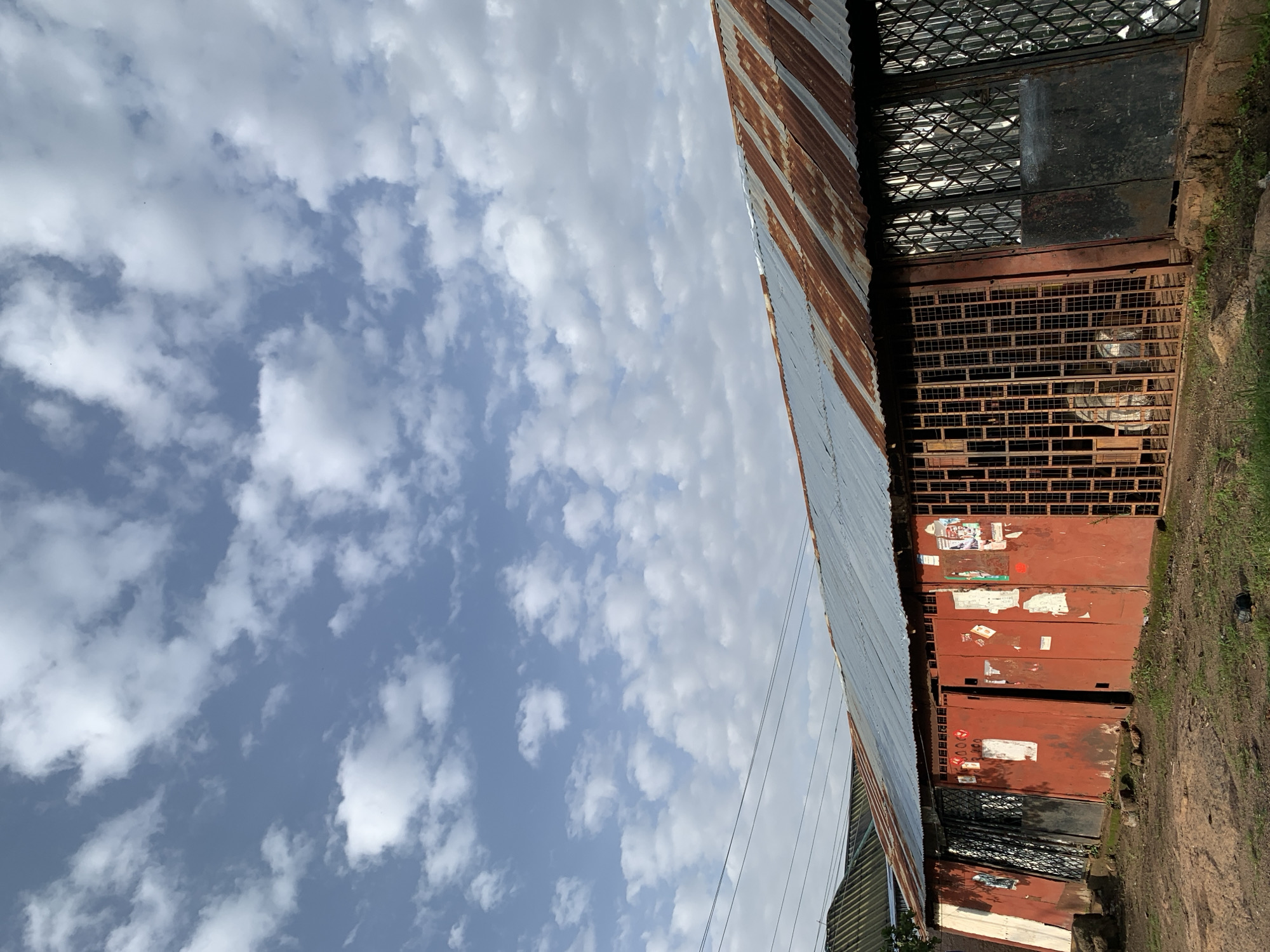 The image depicts a weathered, red-painted building with a corrugated tin roof, exhibiting signs of rust. The building appears to be a shop or a row of shops, with several large metal doors and grilles. Some of these doors are open, revealing glimpses of what might be stored inside, including what appears to be bagged goods. Posters or papers are affixed to some of the doors. The ground in front of the building is unpaved, with patches of dry grass and dirt. Overhead, electrical wires stretch across the frame, and a bright blue sky filled with scattered white clouds dominates the upper portion of the image. This scene is consistent with the visual characteristics of buildings and streetscapes commonly found in parts of Jos, Nigeria, where such structures and materials are prevalent.
