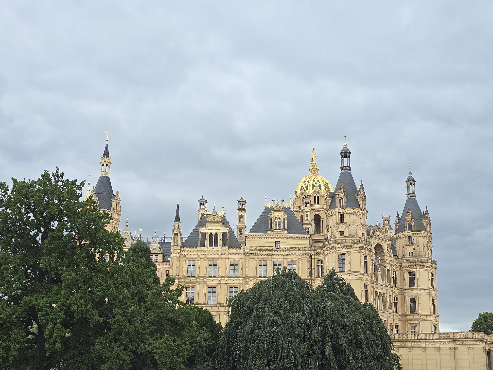 The image shows Schwerin Castle, a picturesque island castle in Schwerin, Germany. The castle is a prominent landmark, with its distinctive architecture and golden domes. It is surrounded by lush greenery, with large trees in the foreground obscuring some of the lower sections of the building. The sky is overcast, suggesting a cloudy day. The castle itself is a grand structure with multiple towers, intricate facades, and numerous windows. The architectural style appears to be a blend of various historical influences, contributing to its unique and imposing presence. The overall scene evokes a sense of historical grandeur and natural beauty.