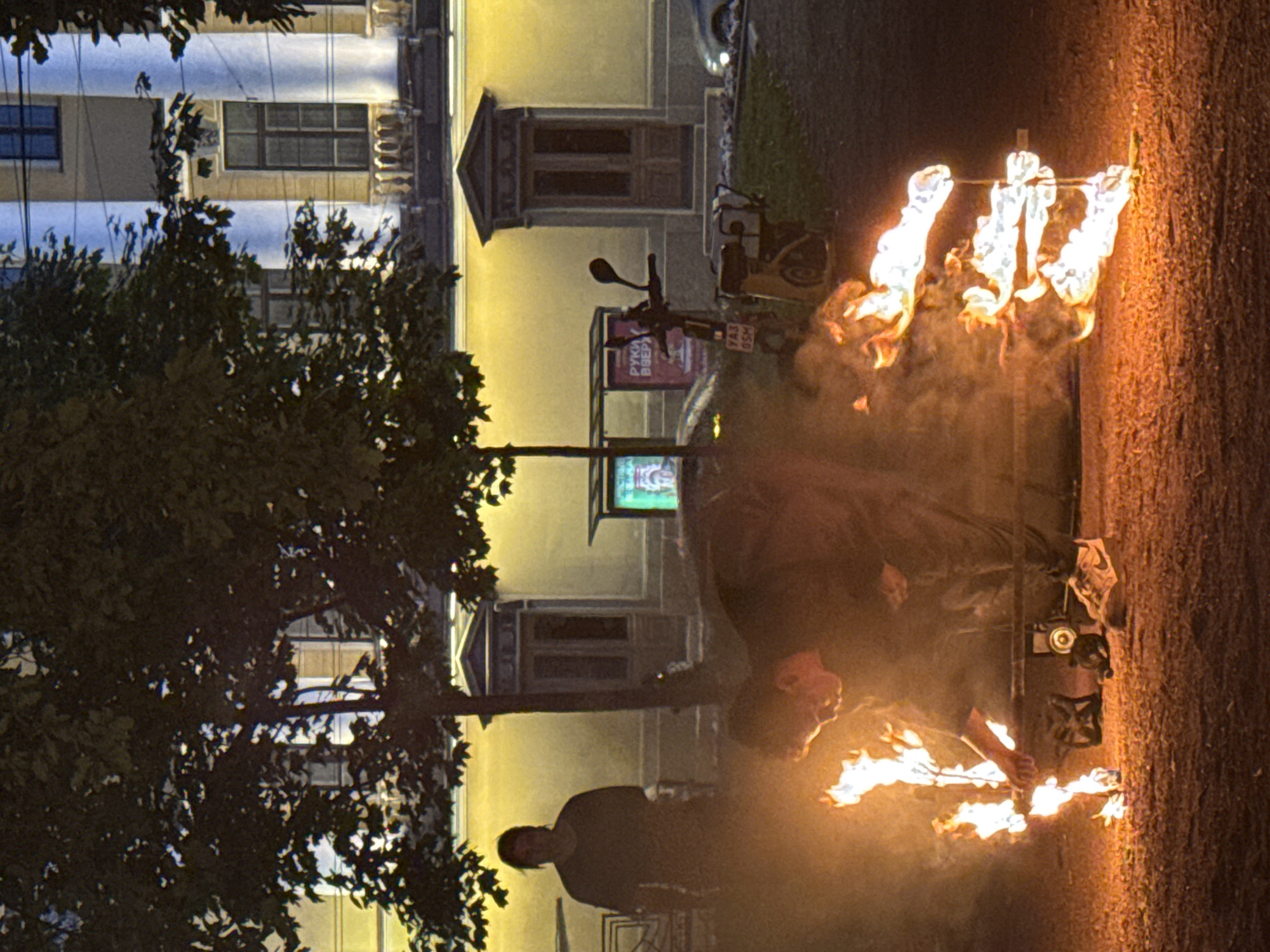 The image captures a street scene at night in Saint Petersburg, Russia, possibly near Palace Square given the architectural style of the building in the background. The primary focus is a person performing with fire. A figure, appearing to be male, is crouched down, holding a burning object that resembles a staff or torch with multiple flaming points. The fire is intense, casting a warm glow on the immediate surroundings and creating smoke.
In the foreground, scattered on the ground, are several objects including what appear to be sneakers and some electronic devices. To the left of the main action, another person stands silhouetted against the light, observing the fire performance.
The background features a building with classical architecture, characterized by large windows, ornate detailing above the doorways, and prominent columns. The building is illuminated by artificial light, creating a yellow hue. A tree with dense foliage is situated in the mid-ground, partially obscuring the view of the building. Various urban elements like a scooter and a sign with Cyrillic text are also visible, suggesting a public urban space. The ground appears to be covered in gravel or small stones.