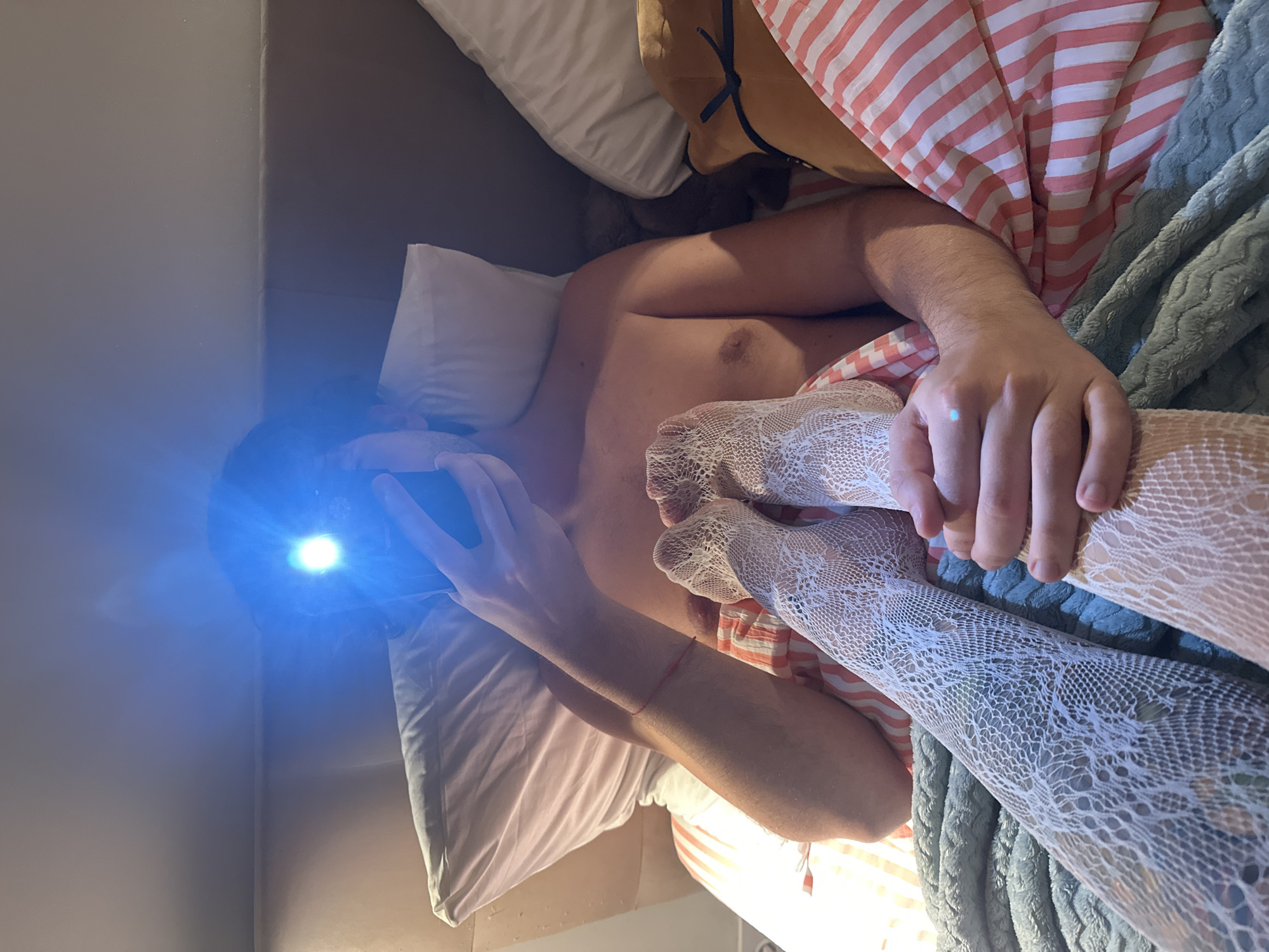 The image shows a person in bed, holding a smartphone with the flashlight on, illuminating their face. The person is wearing white fishnet stockings and is lying on their back. Their legs are crossed. There are pillows and blankets in the background, along with striped bedding. The scene appears to be indoors, possibly a bedroom. There is no information in the image to suggest a specific location within the city of Moscow, Russia.