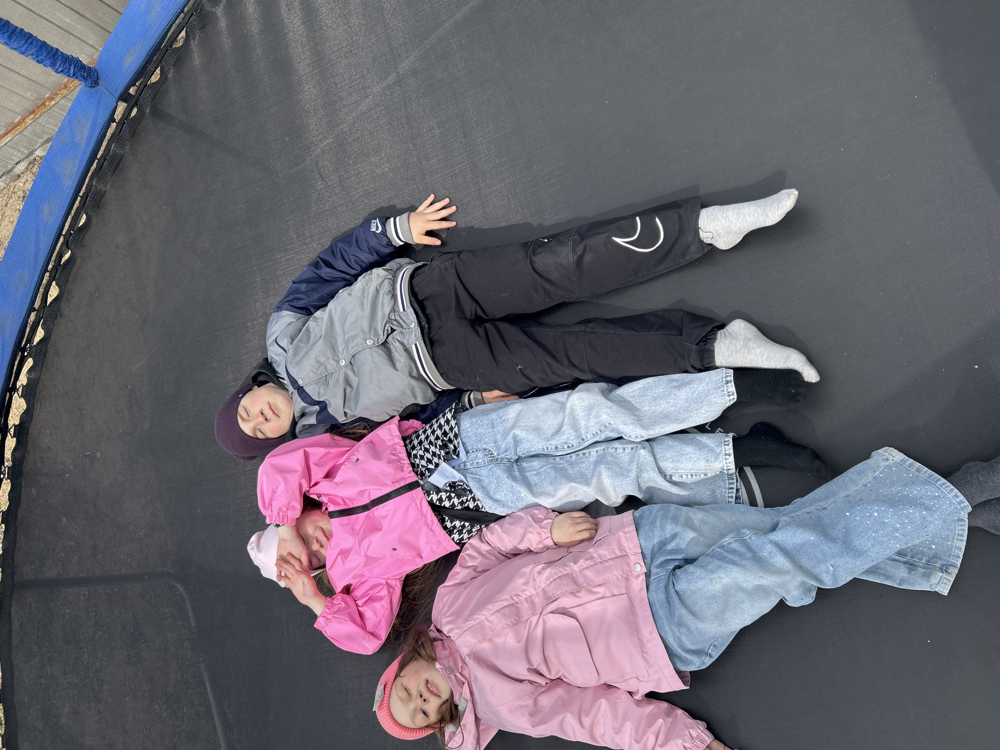 Three children are seen lying side-by-side on a large, black trampoline mat, captured from an overhead perspective. The outdoor setting is brightly lit, indicating daytime and clear weather, and is located in Chaikovsky, Russia.
The child positioned highest in the frame wears a grey and dark blue jacket with a "NIKE" logo on the left sleeve, black pants featuring a white abstract or swoosh-like design, and a dark purple beanie. Their eyes are closed, suggesting rest.
Below, the middle child is dressed in a vibrant pink jacket, a black and white houndstooth top, and light blue ripped jeans. This child's face is slightly turned towards the camera, eyes also closed or relaxed.
The third child, at the bottom of the frame, sports a light pink jacket, light blue jeans adorned with subtle white sparkly dots, and a matching light pink beanie. Their eyes are partially open, conveying a sense of calm.
The trampoline's perimeter is visible with a blue padded edge, and glimpses of the metal frame and springs can be seen beneath. Beyond the trampoline, the background hints at a concrete or dirt ground and a possible fence or structural element. The children appear to be enjoying a relaxed moment, possibly after playing.