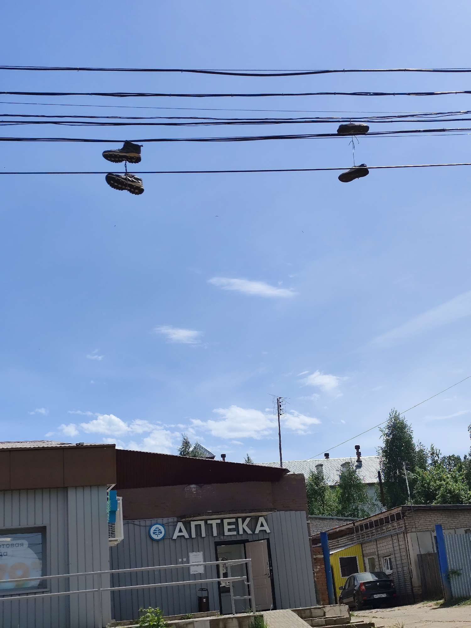 A pair of hiking boots and a pair of flat shoes hang from power lines in the city of Фоки, Russia. The boots are suspended from the lines with their laces, while the flat shoes appear to be tied together and thrown over the lines. Below the power lines is a one-story building with "АПТЕКА" in Cyrillic letters visible on the facade (likely a pharmacy). Further to the right, a small garage-like structure with a dark-colored car parked in front of it is visible. Trees and buildings with white roofs are in the background. The sky is light blue with scattered clouds.