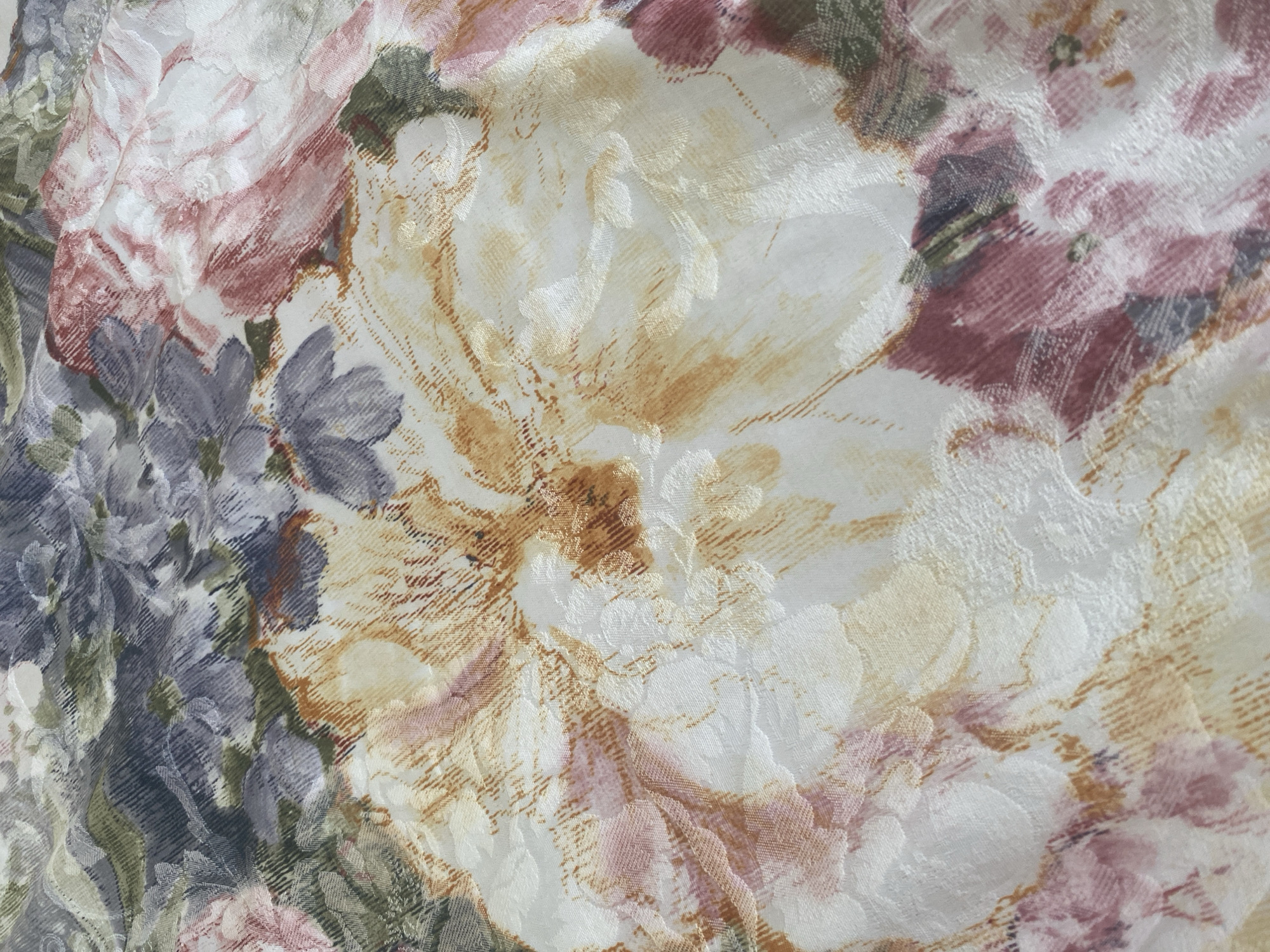 A close-up, overhead shot captures a floral patterned fabric with a satin sheen. The pattern features large, stylized flowers in shades of cream, pale yellow, dusty rose, deep pink, and muted purple, interspersed with green and gray foliage. The fabric is textured, with the floral motifs appearing embossed or woven into the material. The lighting casts subtle shadows, highlighting the raised patterns and creating a sense of depth. No people, animals, or environmental context are visible.