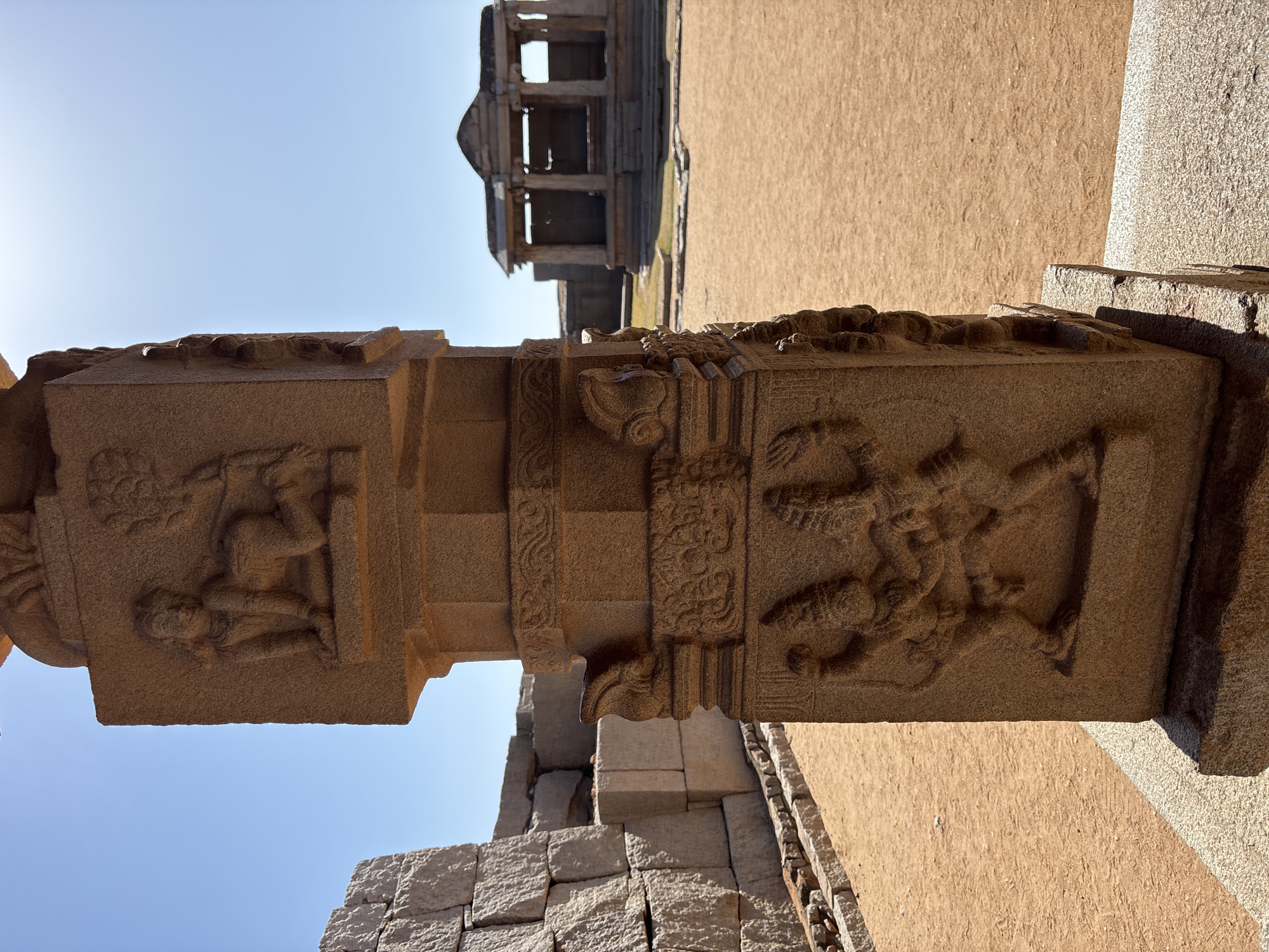 The image shows a detailed stone pillar with carved figures in Kamalapura, India, located in the ruins of Hampi. The pillar is made of a light brown stone and features intricate carvings of human figures in various poses. In the background is a partially destroyed building with multiple levels and pillars, against a clear blue sky. The ground is covered with a layer of sand and stone.