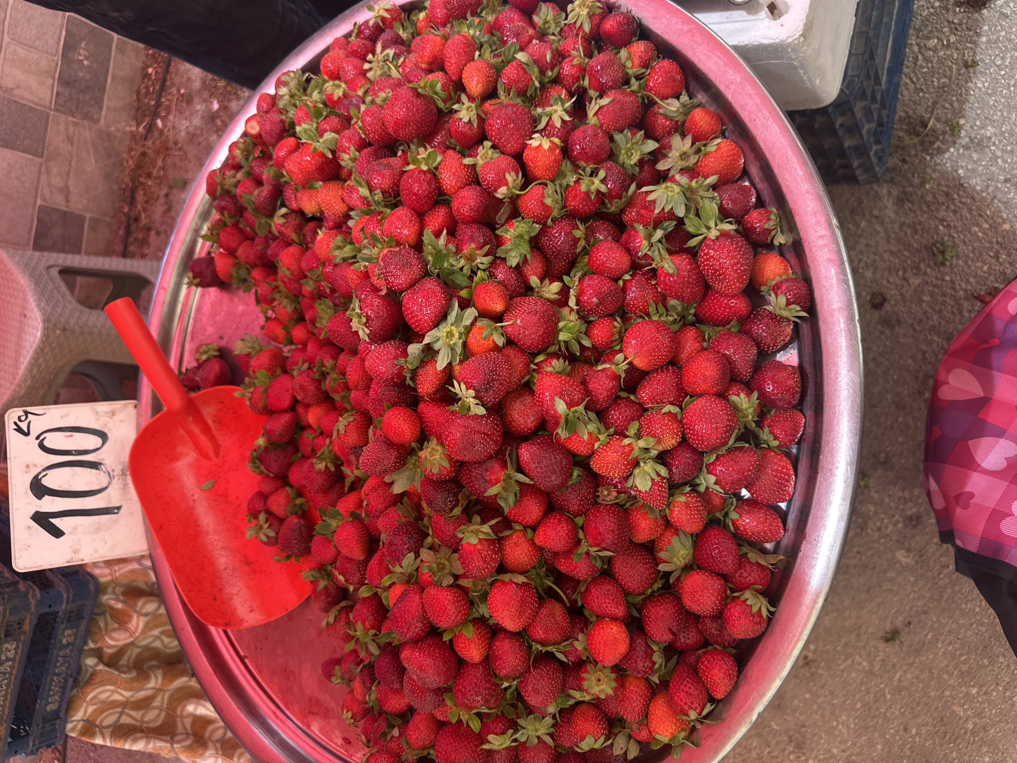 A close-up, high-angle shot captures a large, metallic bowl overflowing with ripe, red strawberries. A bright red plastic scoop rests on top of the strawberries, angled towards the left side of the bowl. A white sign with "100" in black handwritten numbers and a smaller "kg" in the upper right corner is positioned behind the scoop. In the background, blue plastic crates are visible, along with a patterned fabric and a blurry grey and white tiled surface. The overall impression is of a market stall display of fresh fruit.