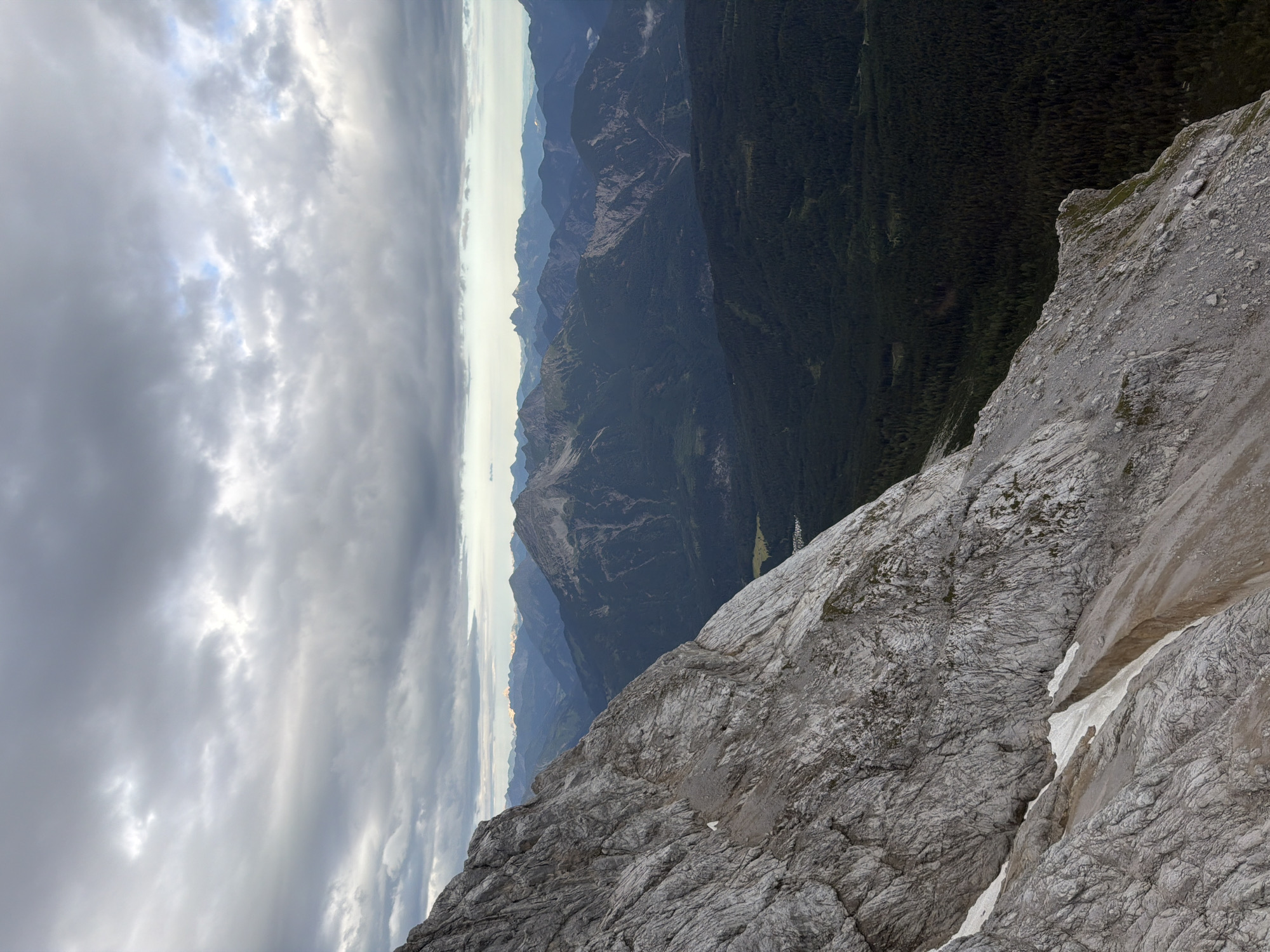 The image depicts a vast mountain landscape, likely taken from a high vantage point in or near Grainau, Germany, given the distinctive alpine scenery. In the foreground, a massive, rugged, and weathered grey rock face dominates the left side of the frame. This rock face is textured with intricate cracks and fissures, showing signs of erosion and age. Patches of snow are visible clinging to the lower sections of the rock face, suggesting a high altitude or recent snowfall.
Beyond the immediate rock face, the terrain drops sharply into a densely forested valley. The forest is predominantly dark green, indicating a thick growth of coniferous trees. This dark expanse of trees stretches into the mid-ground.
Further in the distance, a series of layered mountain ranges recedes towards the horizon. These ranges vary in color from deep blue and grey to lighter shades, with some peaks showing hints of snow or mist. The farthest peaks are softened by atmospheric haze.
The sky above is filled with a dramatic, overcast cloud cover. Large, textured grey clouds dominate the upper portion of the image, with some areas showing lighter patches where sunlight might be trying to break through, or where the clouds are thinner. The overall mood is somber and majestic, characteristic of a cloudy day in the Alps. No people, animals, or man-made structures are clearly discernible in the image. The scene is natural and wild, emphasizing the raw power and beauty of the mountain environment.