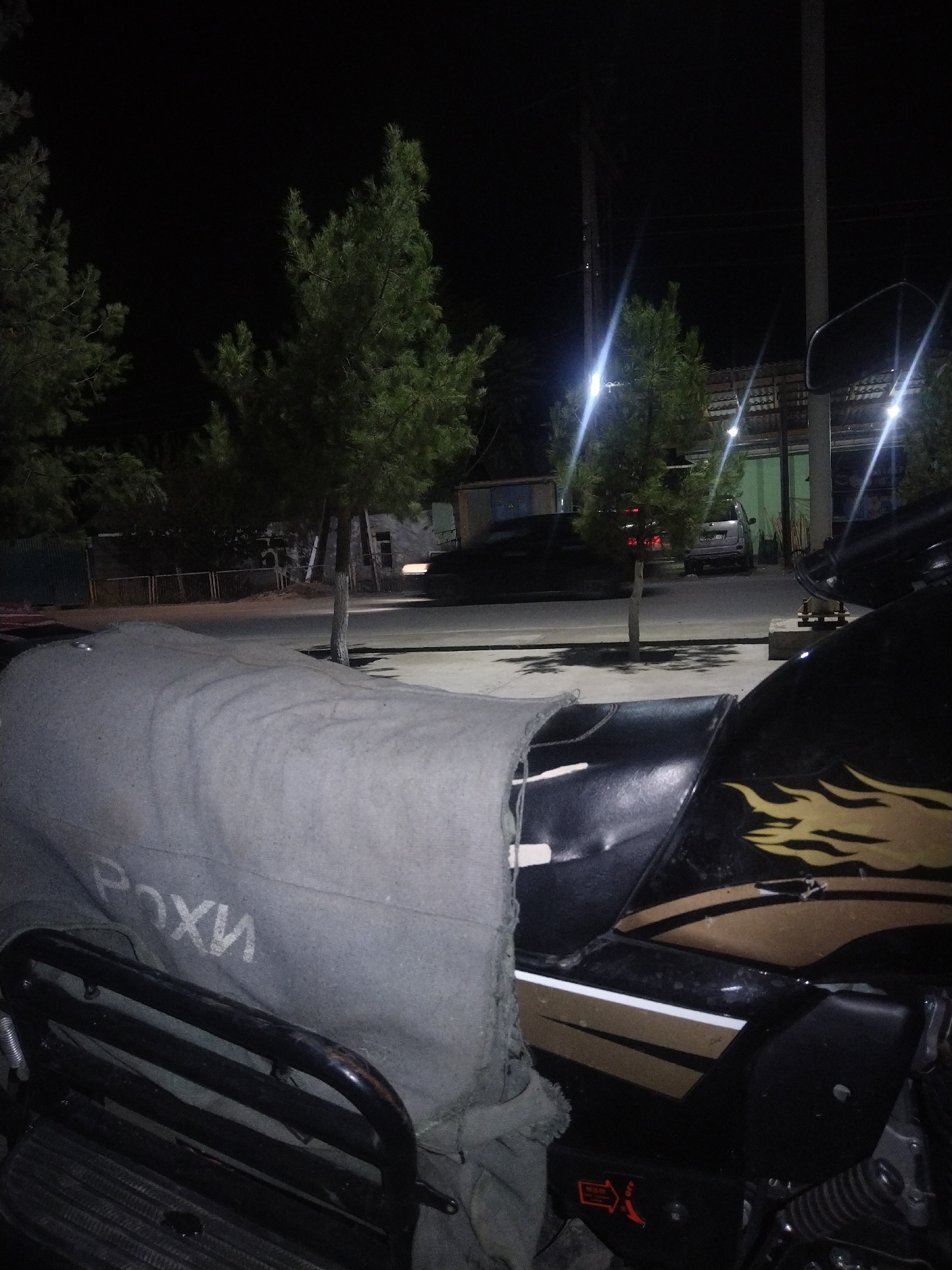 A nighttime outdoor scene shows a motorcycle in the foreground, partially covered by a gray tarp. The tarp has white lettering on it that appears to read "РОХИ". The motorcycle is black with gold flame decals on its side. Behind the motorcycle, a paved area leads to a road with trees and streetlights. In the background, a building with a corrugated roof is visible, along with parked cars. The scene is set in the city of Safarmat, Tajikistan.