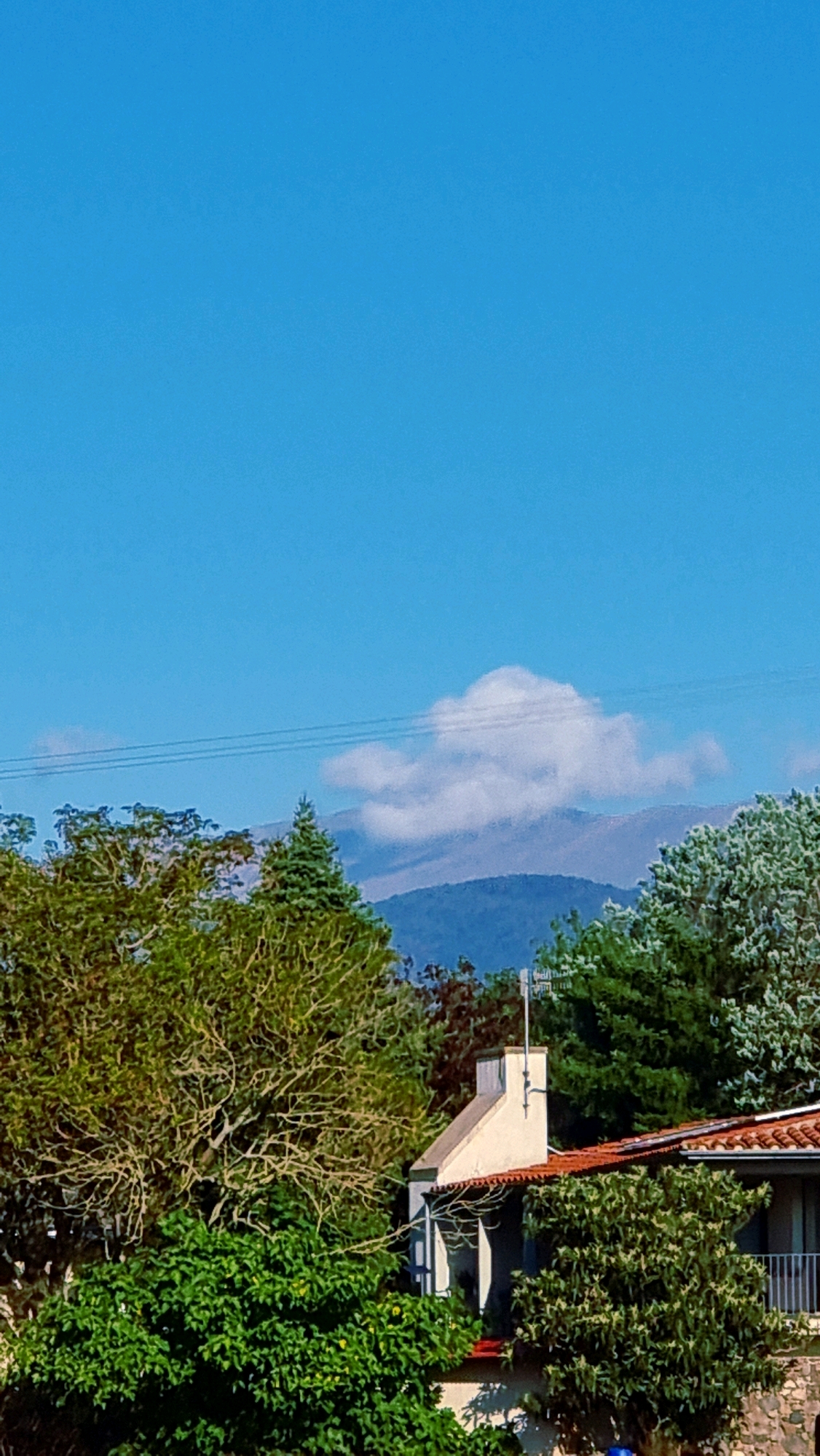 The image depicts a residential scene under a clear blue sky with scattered clouds. In the foreground, lush green trees and vegetation partially obscure a light-colored house with a terracotta tile roof. A chimney with an antenna rises from the roof. In the midground, a dense evergreen tree stands out amidst the other foliage. The background features rolling, blue-toned mountains, capped by a wispy white cloud formation. Power lines are visible stretching across the sky above the trees and mountains. The scene is an outdoor view from a residential area, possibly Llocalou, Spain, given the architectural style of the house and the mountainous landscape. There are no people visible in the image.