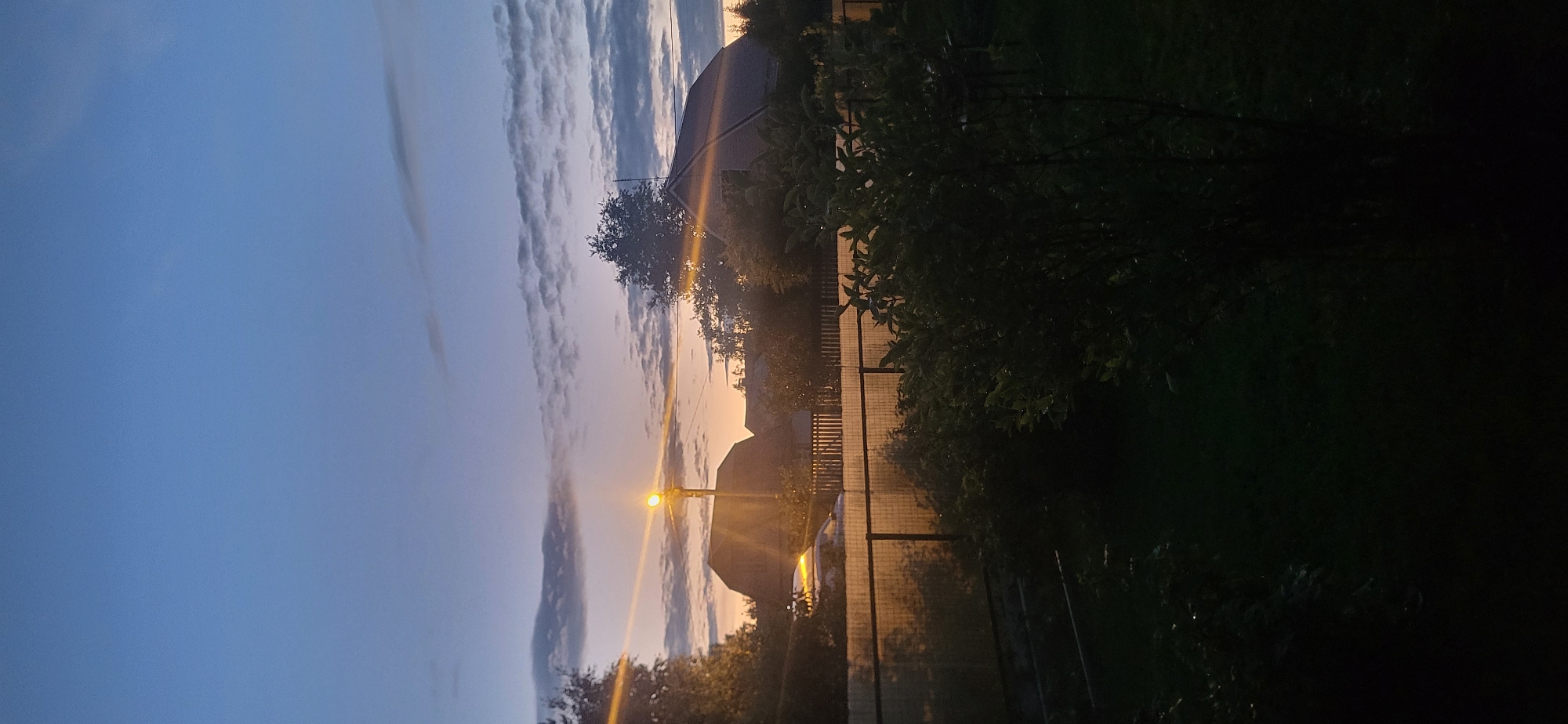 The image depicts a twilight scene in the village of Chaika SNT, Russia. The sky is a gradient of deep blue at the top transitioning to a soft orange and pink hue near the horizon, indicating sunset or sunrise. Wispy, thin clouds are scattered across the sky, catching the fading light.
In the mid-ground, several residential buildings with dark roofs are visible. A street lamp with a glowing yellow light stands prominently on a pole, casting rays that spread outwards, creating a lens flare effect. A fence, possibly made of metal or wire mesh with a semi-transparent covering, stretches across the middle of the image, separating the foreground from the background. Beyond the fence, a car with its headlights on is parked.
The foreground is dominated by dark, dense foliage, likely bushes and trees, with a hint of a lawn or ground cover visible in the lower section. The overall lighting suggests it is either early morning or late evening, with the natural light diminishing and artificial light from the street lamp becoming more apparent. No people are visible in the scene. The environment appears to be a rural or semi-rural residential area.