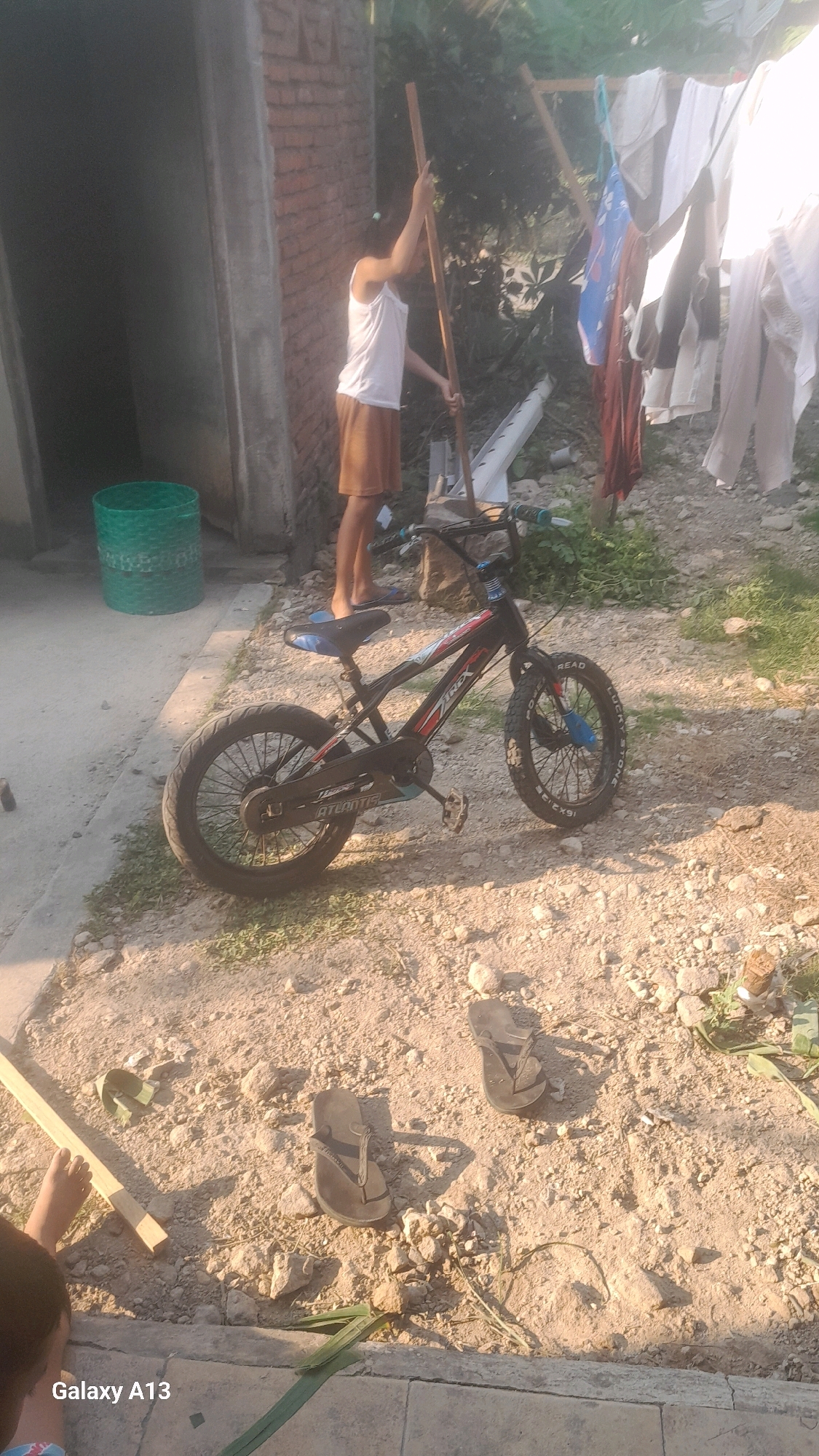 The image depicts an outdoor scene, likely in a rural or semi-urban area in Ngelo, Indonesia. A young person, wearing a white tank top and brown shorts, is standing next to a brick wall and holding a long wooden pole. They appear to be interacting with laundry hanging on a makeshift clothesline. A green woven basket is visible on the ground to the left of the person. A black bicycle with blue accents is parked in the foreground. The ground is unpaved, consisting of dirt and gravel, with some scattered rocks and sparse vegetation. In the lower right corner of the frame, a pair of brown flip-flops are visible on the ground. The overall impression is one of everyday life and outdoor chores.
