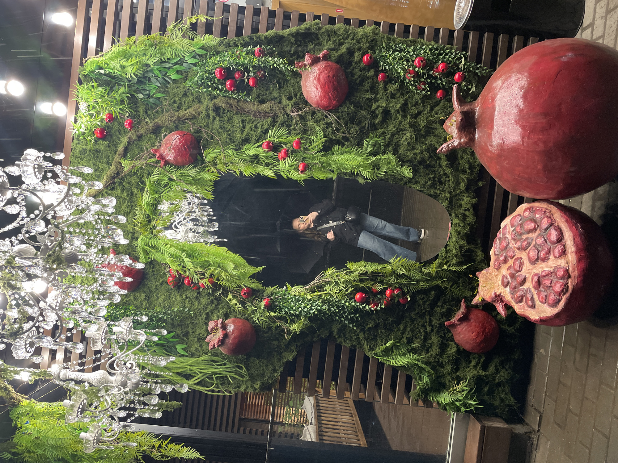 The image shows a decorative display in an urban environment. The prominent feature is a moss-covered structure adorned with faux greenery and pomegranate-shaped decorations of varying sizes. A large, ornate chandelier is positioned to the left of the structure. A reflective surface is within the moss-covered display, and it is reflecting a person wearing a black jacket and blue jeans. The person is holding a phone. The background consists of wooden slats and what appears to be an exterior brick-like surface. The location is Izhevsk, Russia, based on information in search queries.