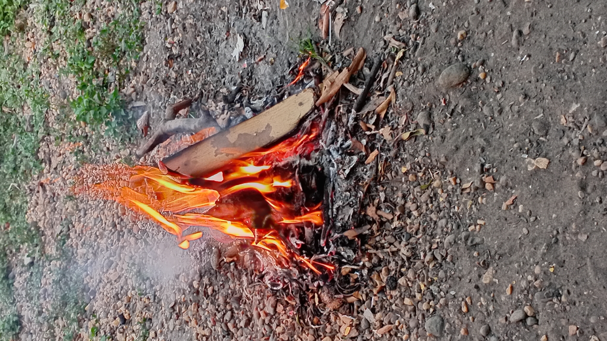 A small campfire burns in a stony, outdoor area with some green vegetation visible in the background. The fire is contained within a pile of small stones, ash, and dried leaves. A piece of wood is positioned within the flames, and smoke rises from the fire. The ground is covered in a mix of dirt and small, gray and tan-colored pebbles. The scene appears to be in Kasang Pudak, Indonesia.