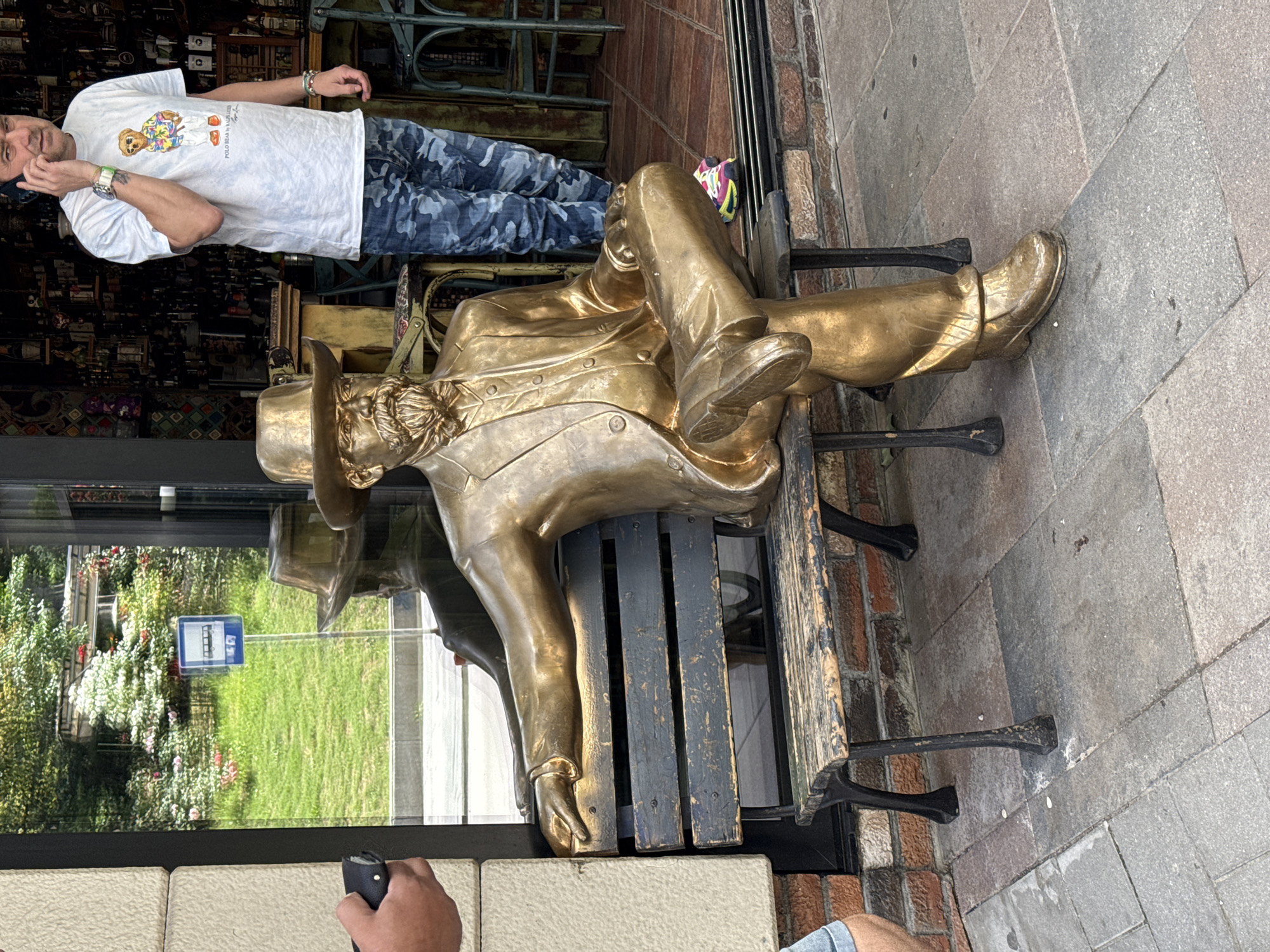 Here's a detailed description of the image:
**Overall Scene:**
The image shows a bronze statue of a man in period attire (likely from the 19th or early 20th century) reclining on a bench outside. The background suggests an urban setting, possibly a shopping or tourist area. A man stands nearby, engrossed in a cell phone conversation.
**Statue Details:**
* **Material:** Bronze or a bronze-colored metal.
* **Attire:** The figure is dressed in a suit, including a jacket, trousers, and shoes.
* **Pose:** The statue is posed in a relaxed, reclining position, as if resting on the bench.
* **Hat:** The statue also features a hat.
**Surrounding Elements:**
* **Bench:** The statue rests on a wooden bench with metal armrests and legs.
* **Building/Shop Front:** Behind the bench is the facade of a building or shop, with glass windows and some kind of structural support.
* **People:** A man wearing a white T-shirt is talking on a cell phone nearby.
* **Pavement:** The scene is on a paved area, likely a sidewalk or pedestrian zone.
* **Reflection:** Through the glass, a reflection of green vegetation and a street sign can be seen.
**Context:**
Without explicit confirmation, the image's location being the city of Veliko Tarnovo, Bulgaria, relies solely on external information.