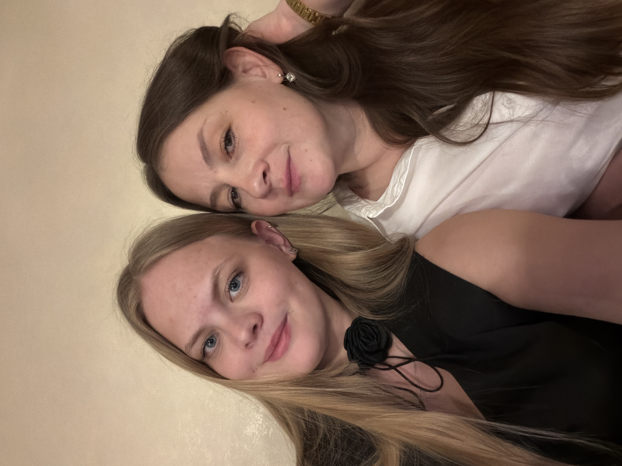 Two women are closely positioned side-by-side, with the blonde woman in the foreground and the brunette woman slightly behind. The blonde woman has light blonde hair, blue eyes, and a light complexion. She is wearing a black top adorned with a dark rose detail. The brunette woman has brown hair, darker eyes, and is wearing a white shirt. Her hand, with a gold watch, is raised towards her hair. The background is a neutral, light wall. There is no specific information to conclude the location is in Izhevsk, Russia. The scene seems to be indoors.