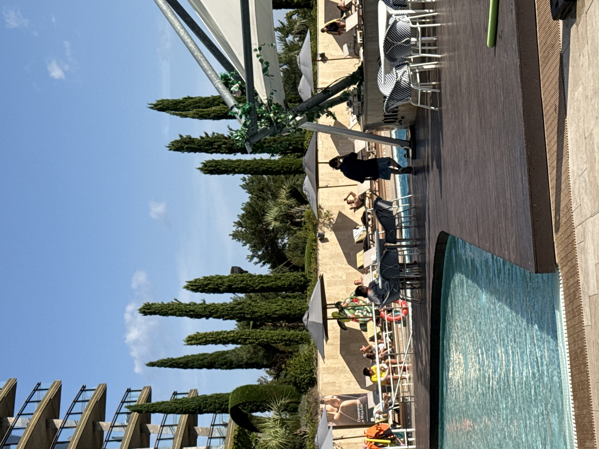 The image depicts a resort pool area on a sunny day. A multi-story building with balconies is visible to the left, partially obscured by tall, slender trees. In the foreground, a swimming pool with clear blue water is bordered by a wooden deck. Several lounge chairs are set up around the pool, some occupied by people. A white canopy structure with metal supports extends over a seating area with tables and chairs. In the background, more trees and vegetation are present, with a clear blue sky overhead. There is no indication in the image that this location is in the city of Симеизский поселковый совет, Ukraine.