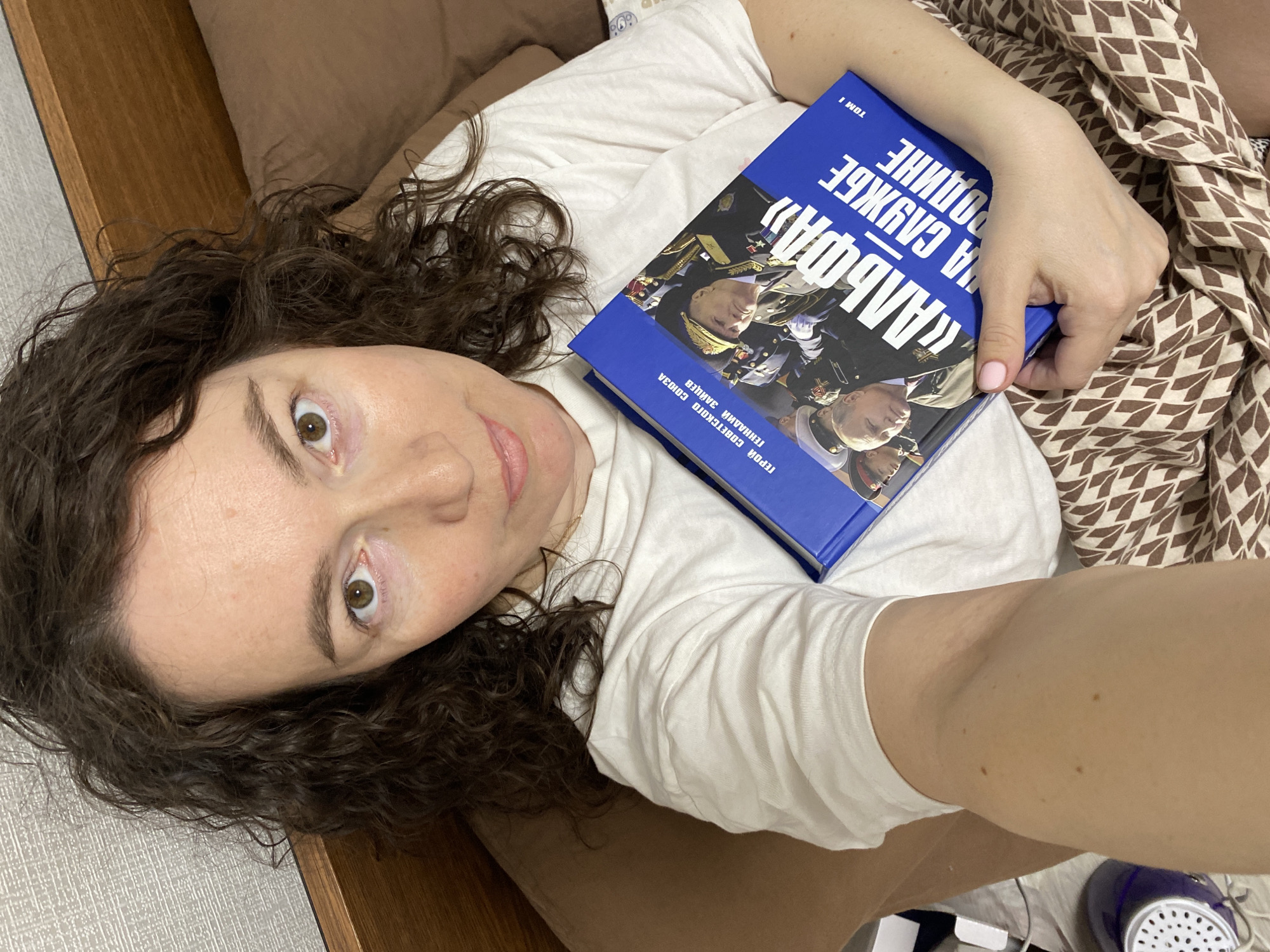 A woman with curly dark hair is lying down, looking at the camera, and holding a blue book titled "Alpha: In the Service of the Motherland" by Gennady Zaitsev, Hero of the Soviet Union. She's wearing a white t-shirt and lying on a bed with a brown headboard and pillow. The bedspread is brown and white. The scene is indoors, possibly in a residential setting. The location of Ольховка, Russia cannot be verified from the image.