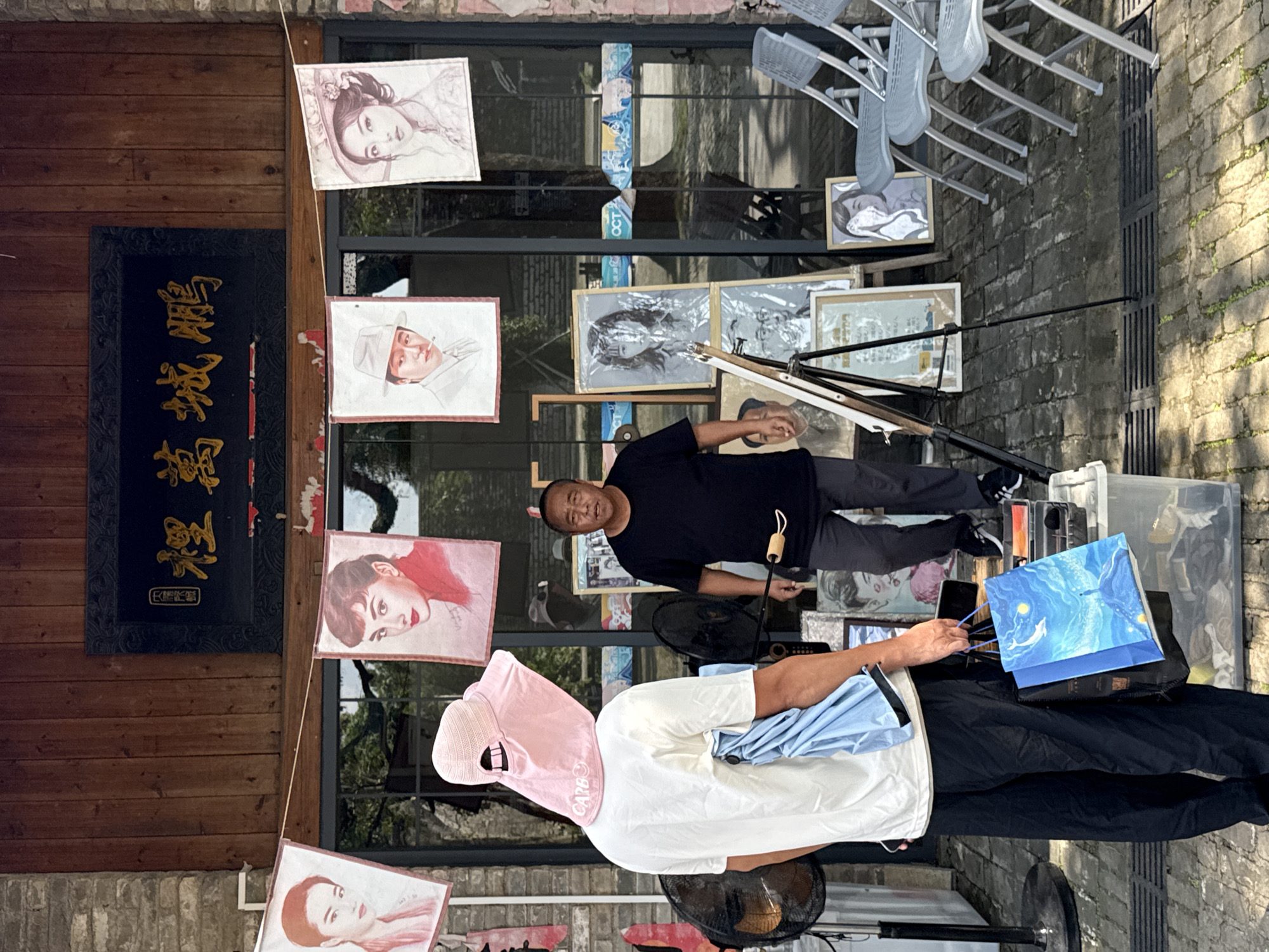 In the city of Longgang District, China, a scene unfolds outside a shop with a wooden facade. A man, wearing a black t-shirt and dark trousers, stands near a display of artwork. To his left, a person in a white t-shirt and dark trousers is wearing a large pink sun hat. They are holding a blue shopping bag adorned with a starry sky and a whale. Several framed portraits and drawings are displayed on easels and leaning against the shop's glass windows. Among these artworks are portraits of Audrey Hepburn and other individuals. A black electric fan is positioned to the left of the person in the pink hat. The ground is paved with bricks, and a metal grate is visible in the foreground. The shop has a sign with Chinese characters in gold against a black background.