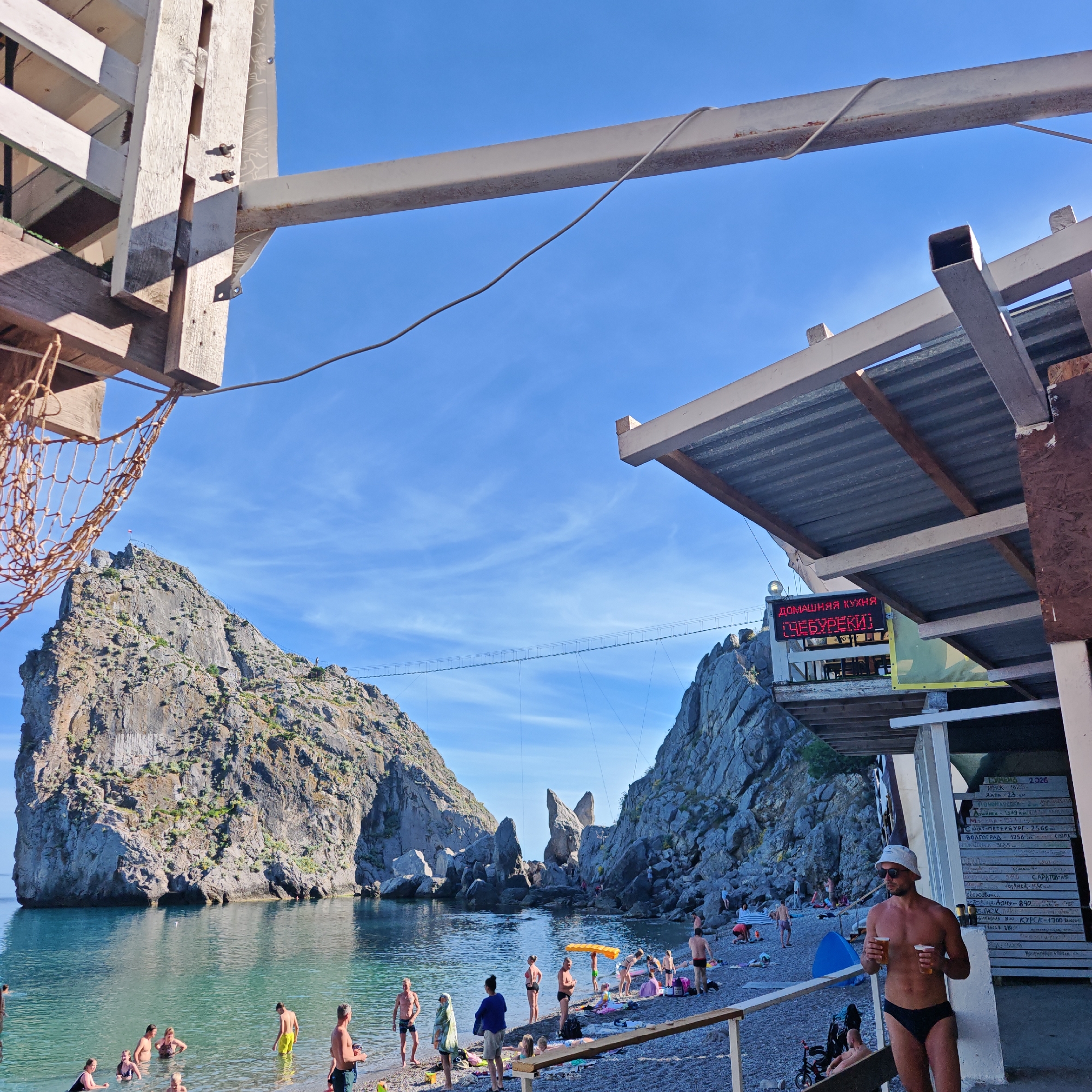 The image depicts a beach scene with a rocky coastline and clear blue water. In the foreground, a man wearing a white hat and sunglasses, and black swim briefs, stands holding two glasses of beer. Behind him, a menu board with Russian text and a digital sign displaying "ДОМАШНЯЯ КУХНЯ ЧЕБУРЕКИ" are visible. The beach is populated with many people, some in the water and others relaxing on the shore. A suspension bridge connects two large rock formations in the distance. The environment appears to be a sunny day with a clear sky.