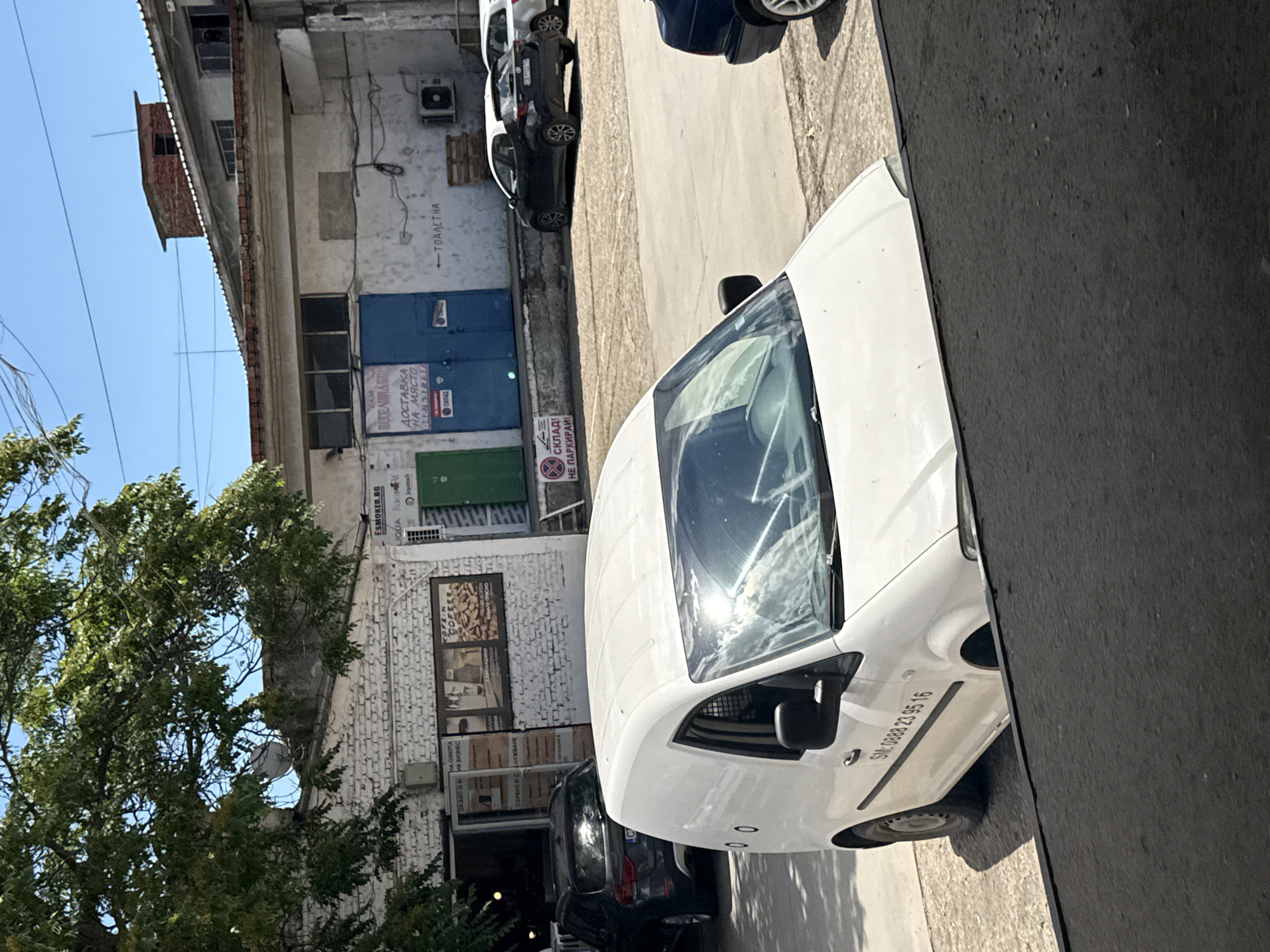 The image depicts a sun-drenched, outdoor scene, likely in an industrial or commercial area of Stara Zagora, Bulgaria. In the foreground, a white panel van with visible smudges and reflections on its windshield is parked. The van has a phone number "SM: 0888 23 95 16" printed on its side. Behind the van, a dark-colored SUV is parked to the left. The background features a weathered, light-colored building with a corrugated metal roof. Signs on the building are visible, including one with "ESMOKER.BG" and another with "ДОСТАВКА НА МЯСТО" (delivery on site). To the right, a black hatchback car and a portion of a white car are parked. A blue door is visible on the building, and further to the right, a sign with a red circle and a diagonal line over a symbol indicates no parking. An outdoor air conditioning unit is mounted on the wall of the building. Green trees with dense foliage are present on the left side of the image, casting shadows. The sky is clear and blue.